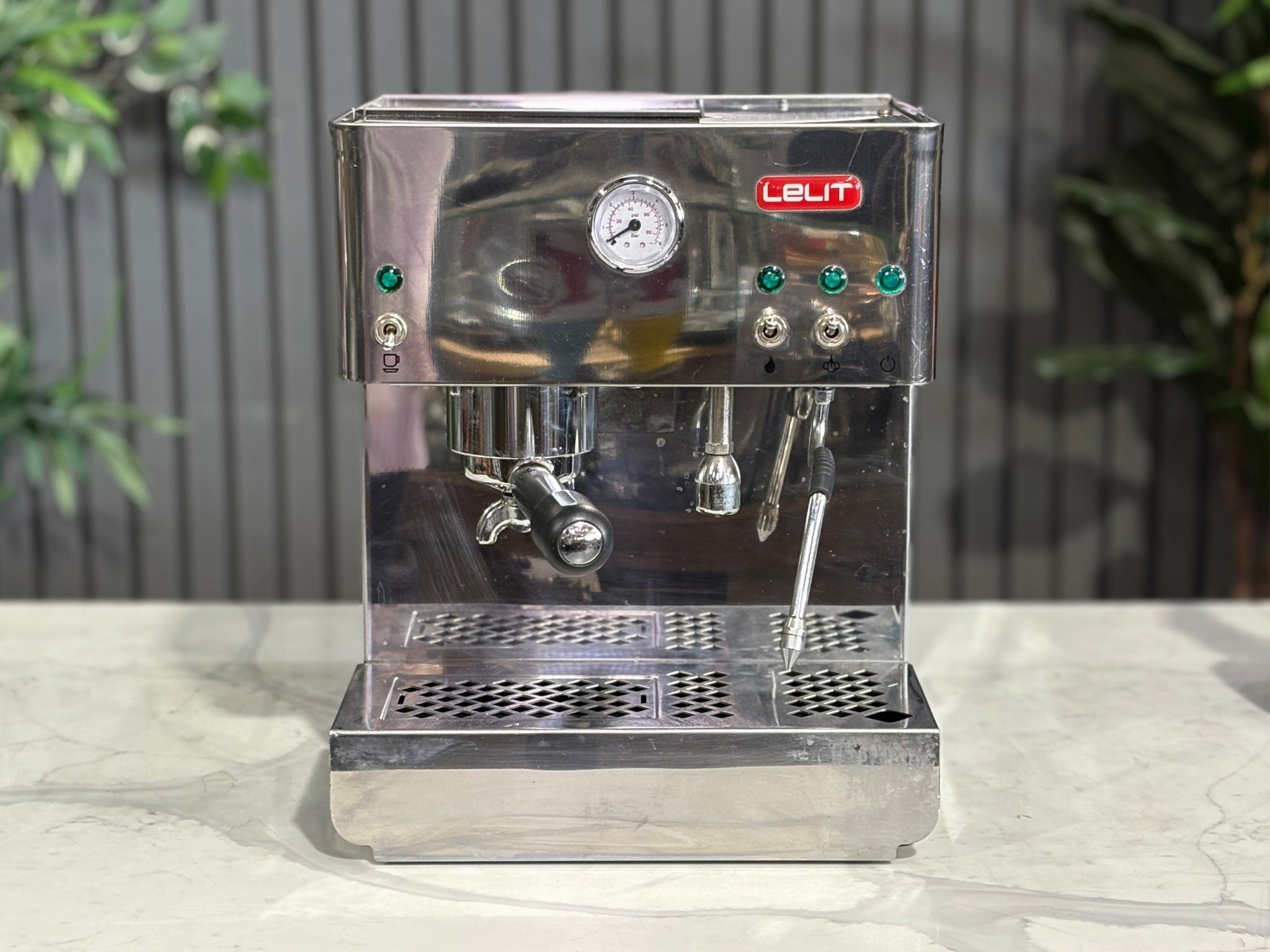 A Lelit Diana PL60 1 Group Coffee Machine with Stainless Steel finish sitting on a marble bench. There is greenery in the background. The image is taken from the front of the machine so that you can see the front of the machine, control panel, buttons and group head.