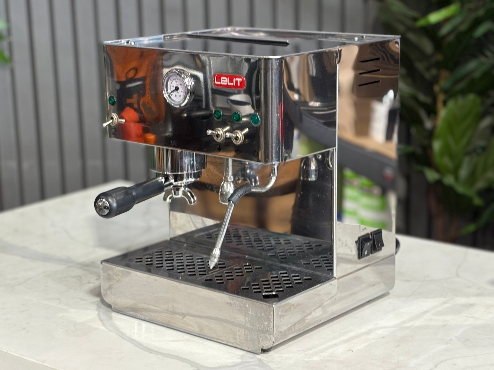 A Lelit Diana PL60 1 Group Coffee Machine with Stainless Steel finish is sitting on a marble bench. There is greenery in the background. The image is taken from the front of the machine and the machine is sitting on an angle so that you can see the front of the machine and the side.