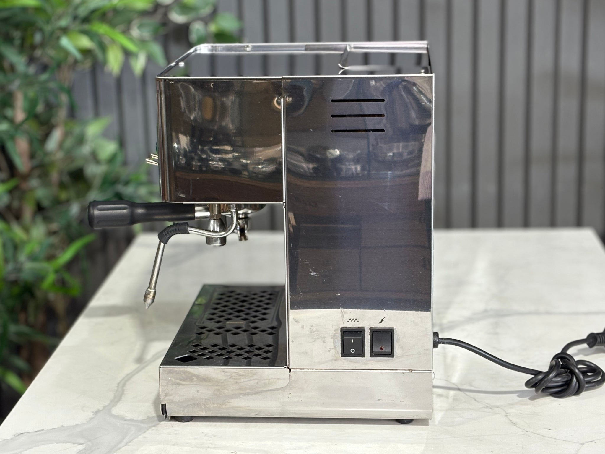 A Lelit Diana PL60 1 Group Coffee Machine with Stainless Steel finish sitting on a marble bench. There is greenery in the background. The image is taken from the side of the machine.