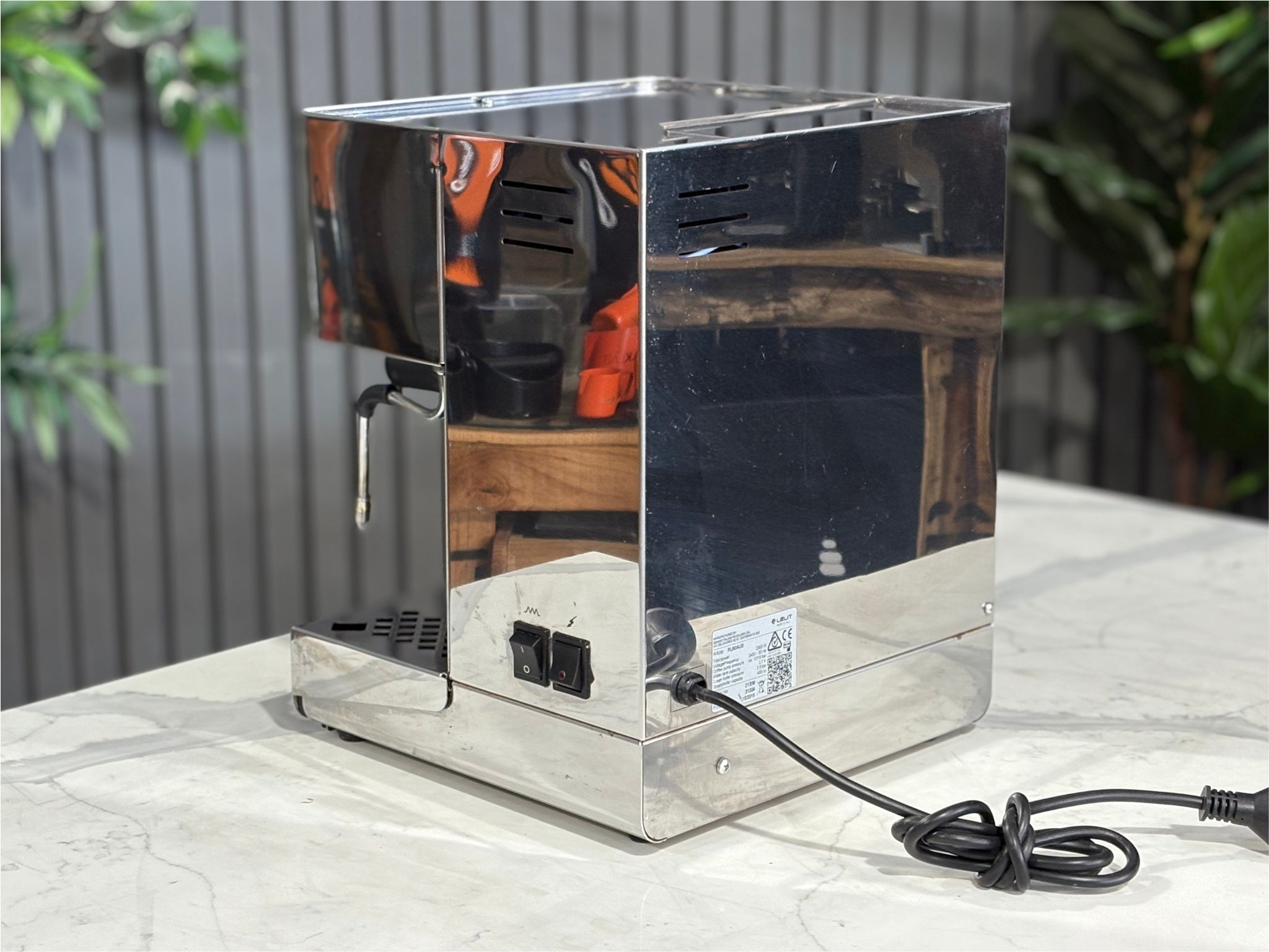 A Lelit Diana PL60 1 Group Coffee Machine with Stainless Steel finish. sitting on a marble bench. There is greenery in the background. The image is taken from behind the machine and the machine is sitting on an angle so that you can see the back of the machine and the side.