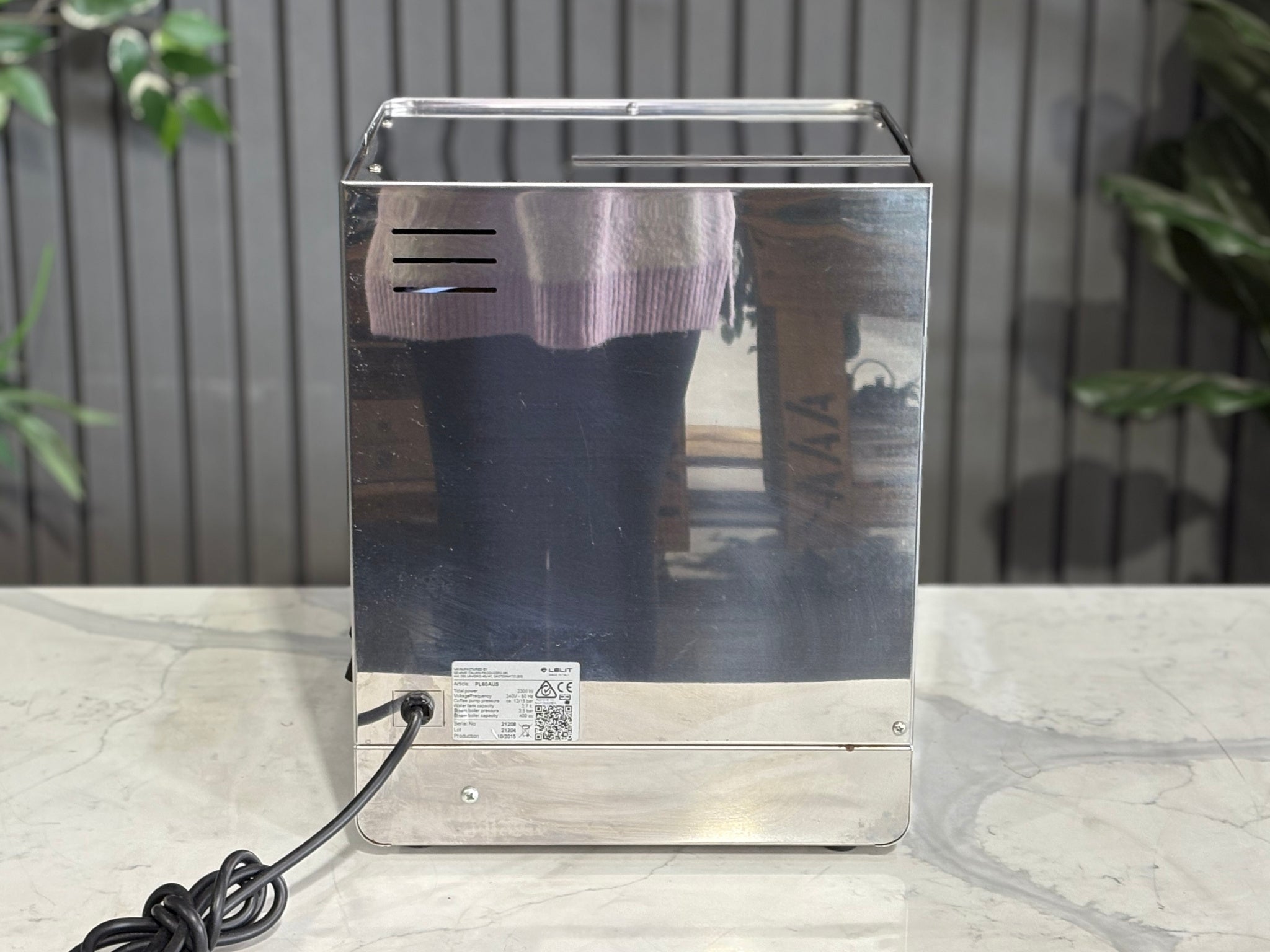 A Lelit Diana PL60 1 Group Coffee Machine with Stainless Steel finish sitting on a marble bench. There is greenery in the background. The image is taken from behind the machine and the machine is sitting on an angle so that you can see the back of the machine and the side.