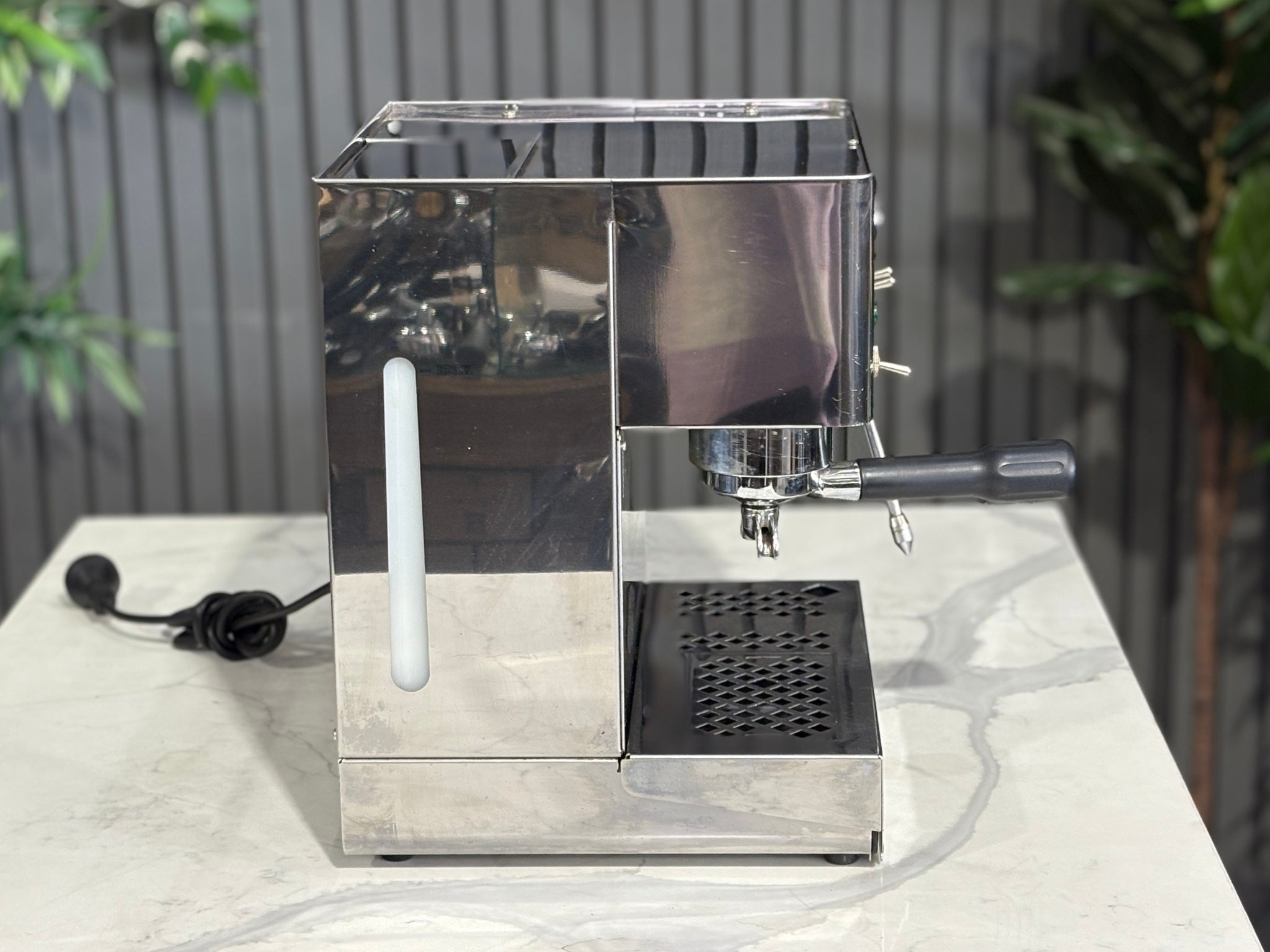 A Lelit Diana PL60 1 Group Coffee Machine with Stainless Steel finish sitting on a marble bench. There is greenery in the background. The image is taken from the side of the machine.