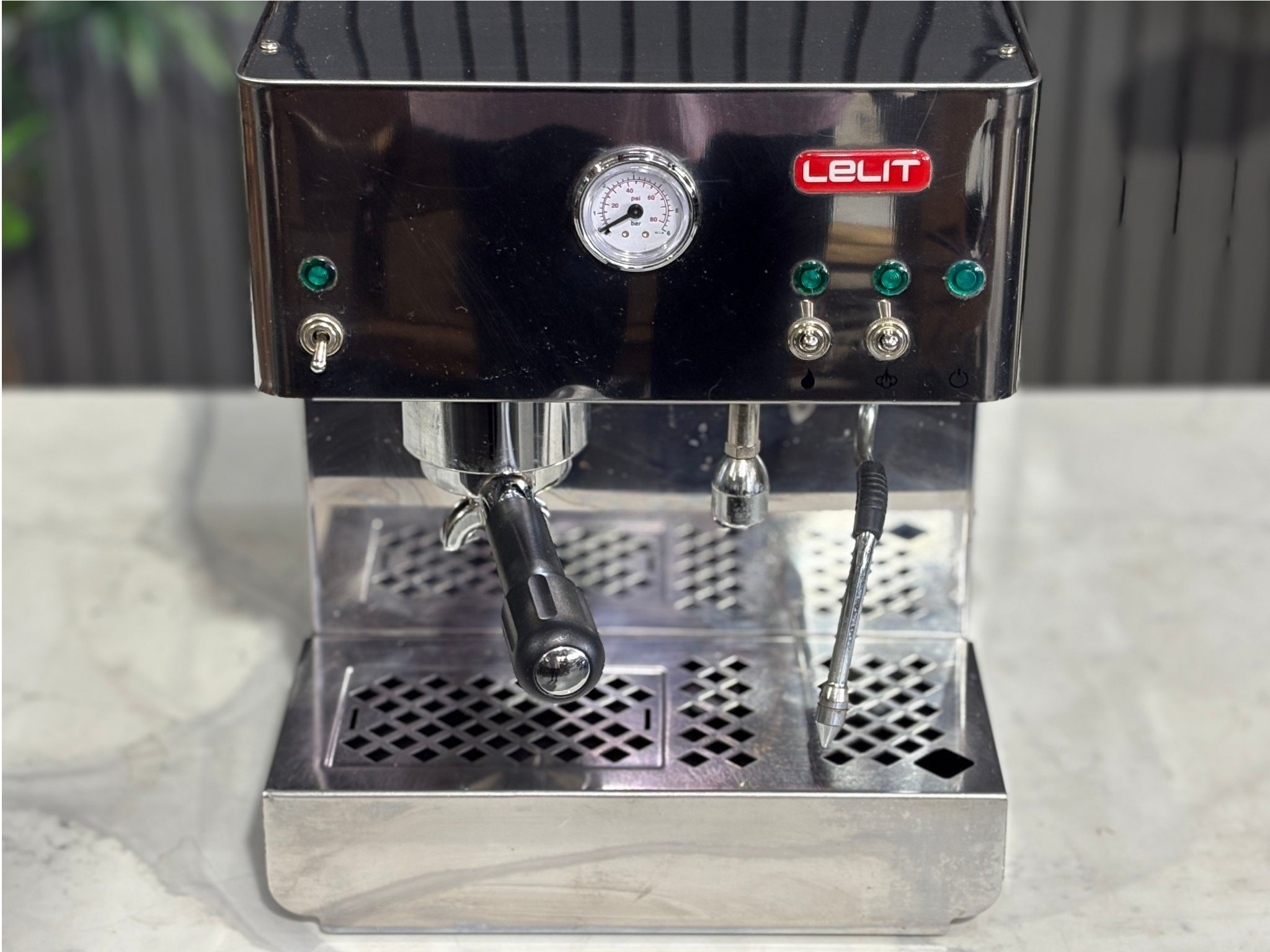A Lelit Diana PL60 1 Group Coffee Machine with Stainless Steel finish
is sitting on a marble bench. There is greenery in the background. The image is a close up image taken from the front of the machine and the machine is sitting on an angle so that you can see the front of the machine, the controls including flick buttons, group handle and logo.