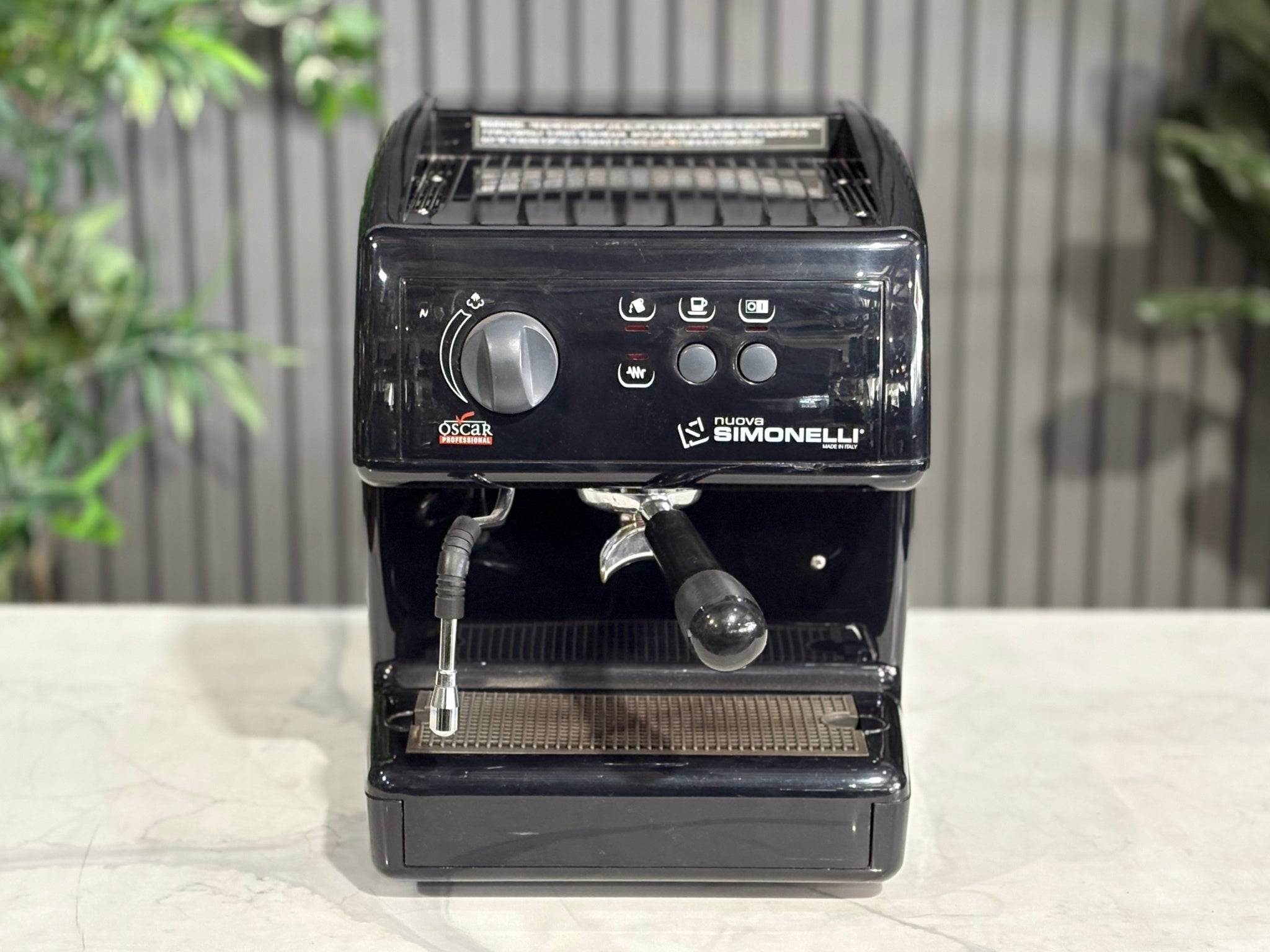 Nuova Simonelli Oscar Professional 1 Group Coffee Machine Black - Used espresso machine photographed from a higher front angle, showing the top panel, cup tray, and sleek black casing. Displayed on a marble bench with greenery behind.