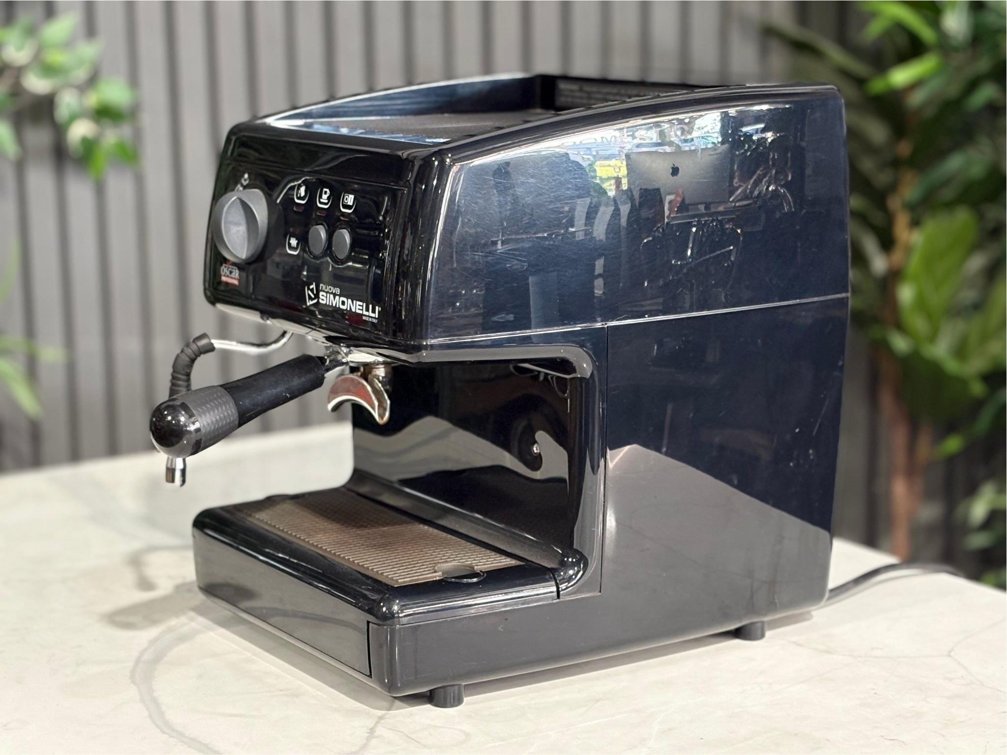 Nuova Simonelli Oscar Professional 1 Group Coffee Machine Black - Used espresso machine displayed at an angle on a marble bench with greenery in the background. Visible features include the button-operated group head, steam wand, and black compact body.