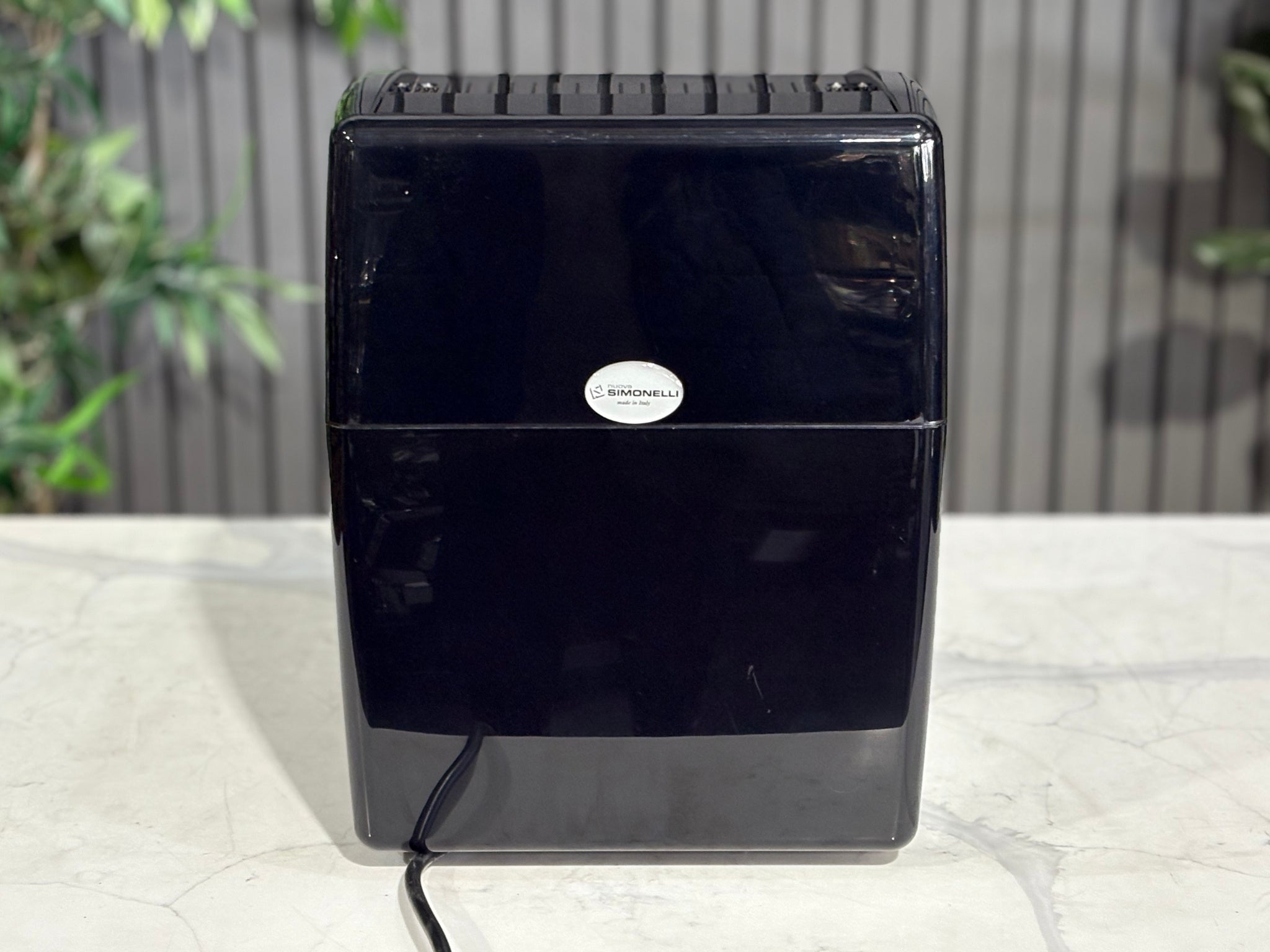 Back view of the Nuova Simonelli Oscar Professional 1 Group Coffee Machine Black - Used espresso machine, revealing the rear panel and streamlined black housing. Set against a marble bench with greenery in the background.