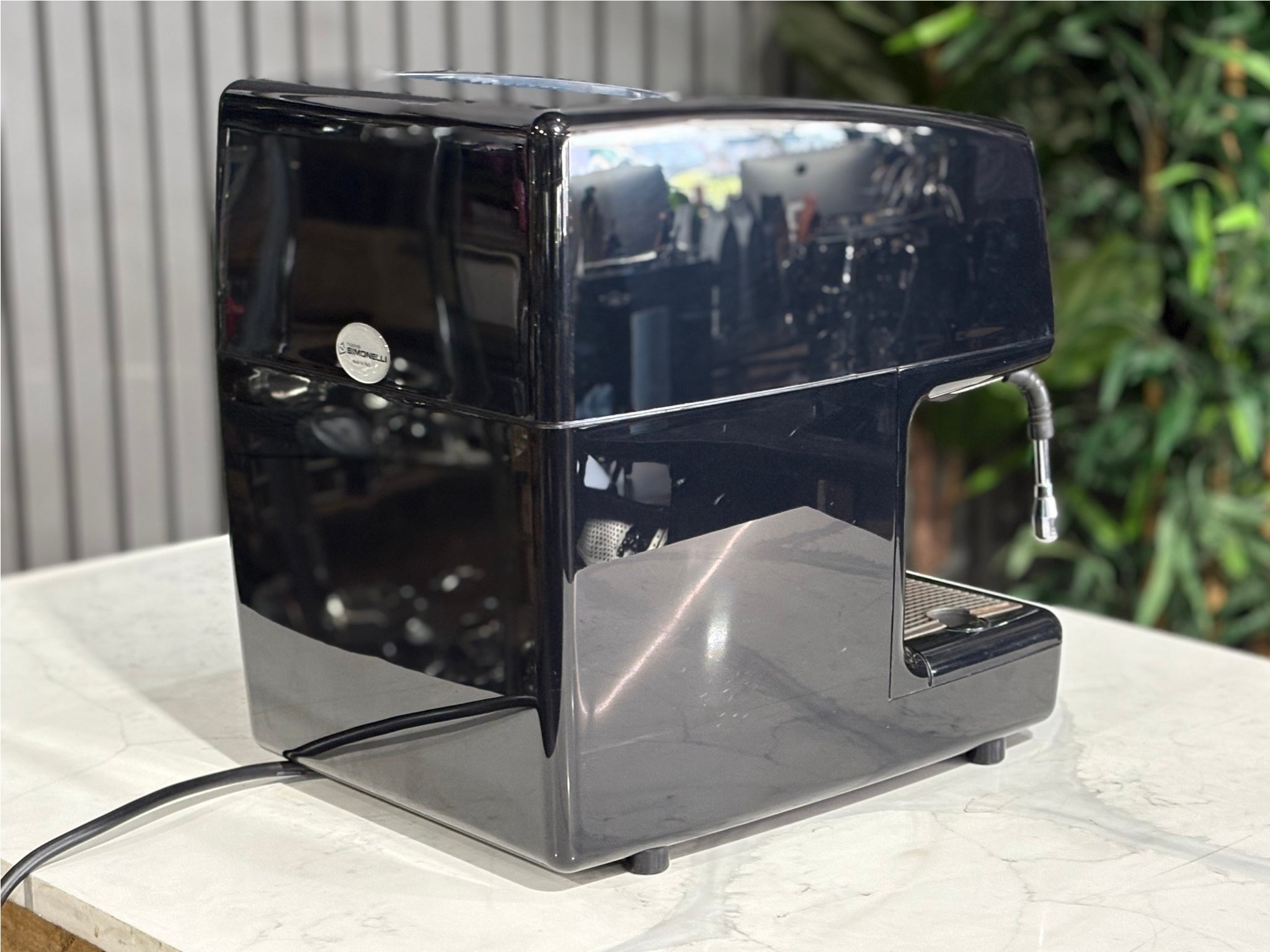 Nuova Simonelli Oscar Professional 1 Group Coffee Machine Black - Used espresso coffee machine viewed from the rear angle, highlighting the body design and black exterior. Displayed on a marble bench with greenery in the background.