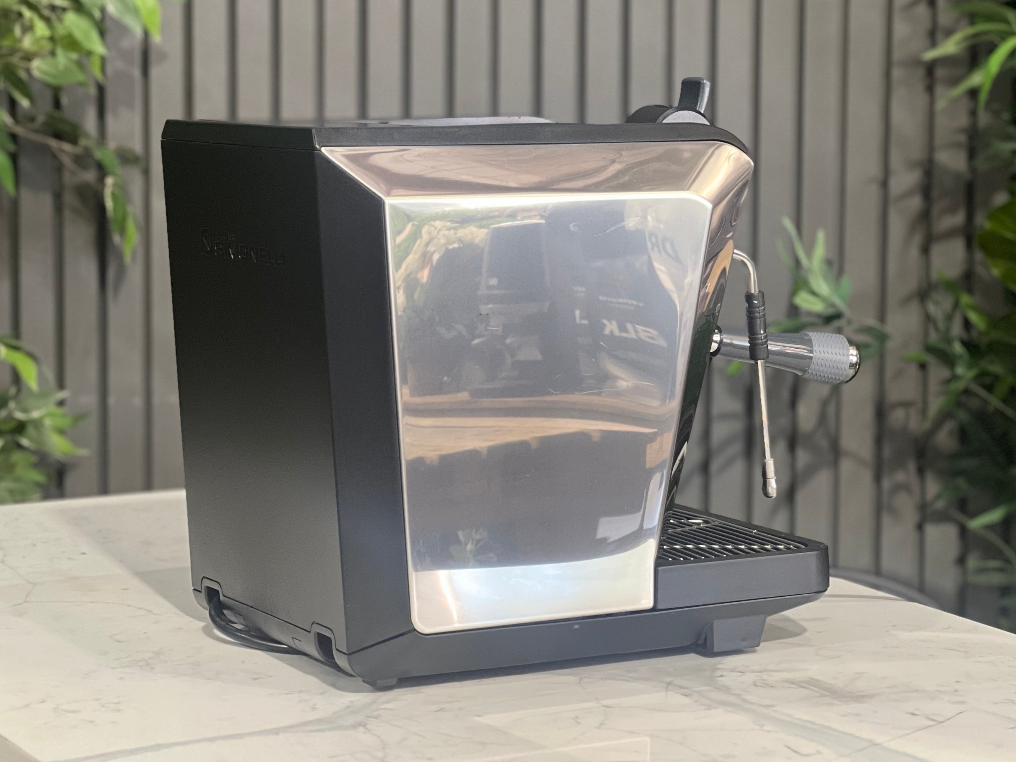 Nuova Simonelli Oscar Professional 1 Group Espresso Coffee Machine Black side view highlighting the full silver side panel, compact form, and integrated water tank housing, styled on a marble bench with lush greenery behind.