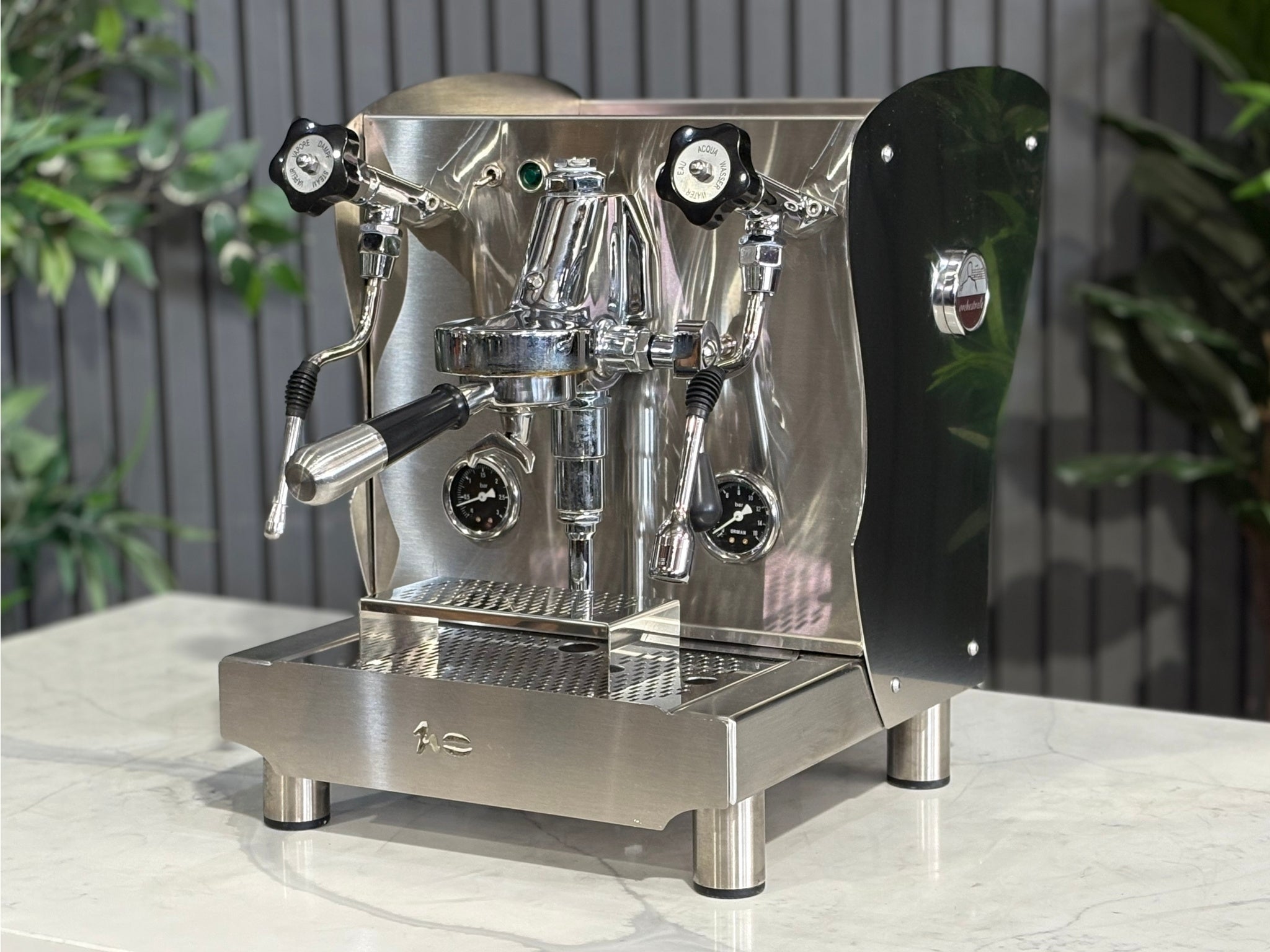 A Orchestrale Nota 1 Group Coffee Machine with Stainless Steel finish is sitting on a marble bench. There is greenery in the background. The image is taken from the front of the machine and the machine is sitting on an angle so that you can see the front of the machine and the side.