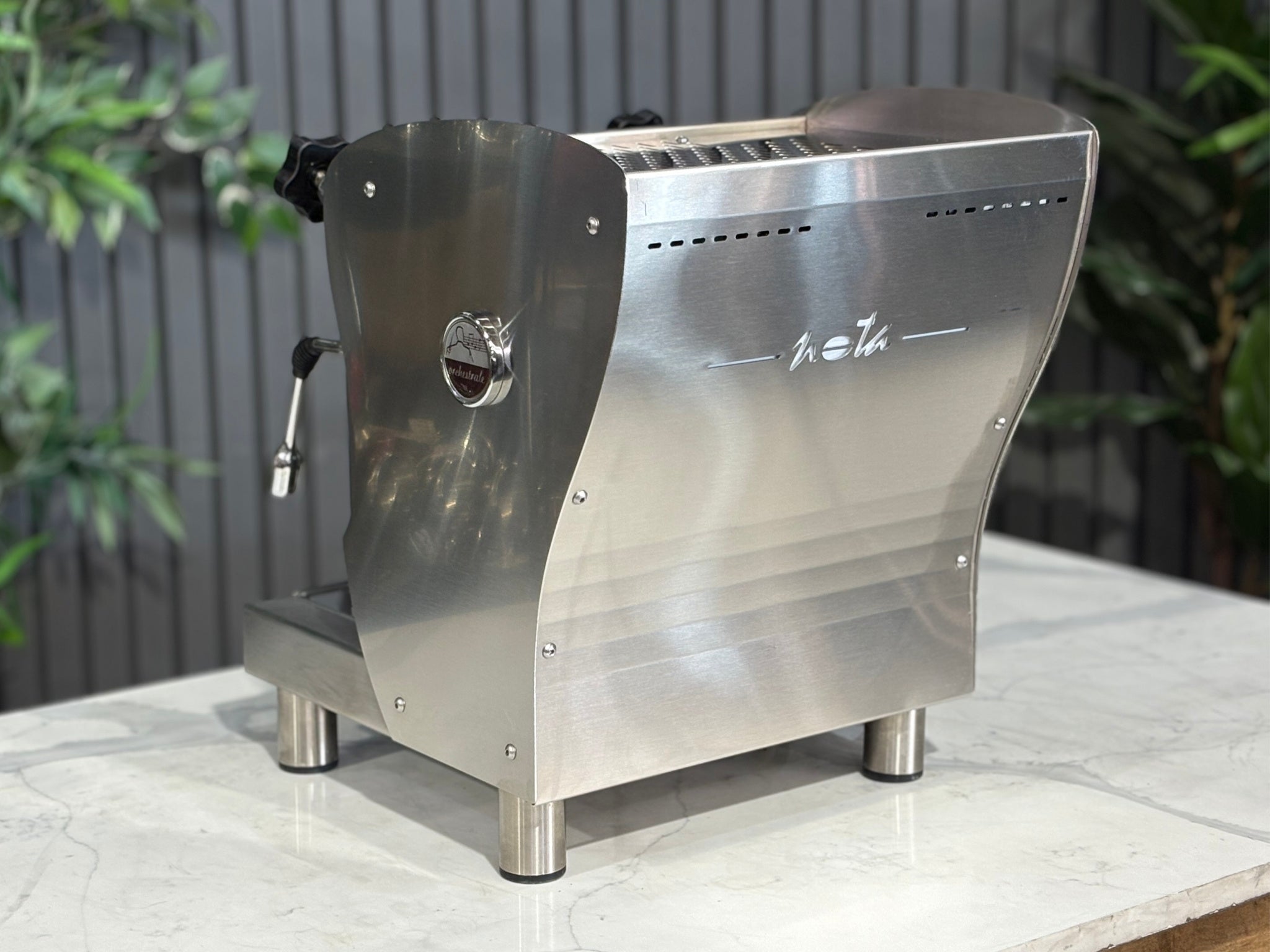 A Orchestrale Nota 1 Group Coffee Machine with Stainless Steel finish sitting on a marble bench. There is greenery in the background. The image is taken from behind the machine and the machine is sitting on an angle so that you can see the back of the machine and the side.