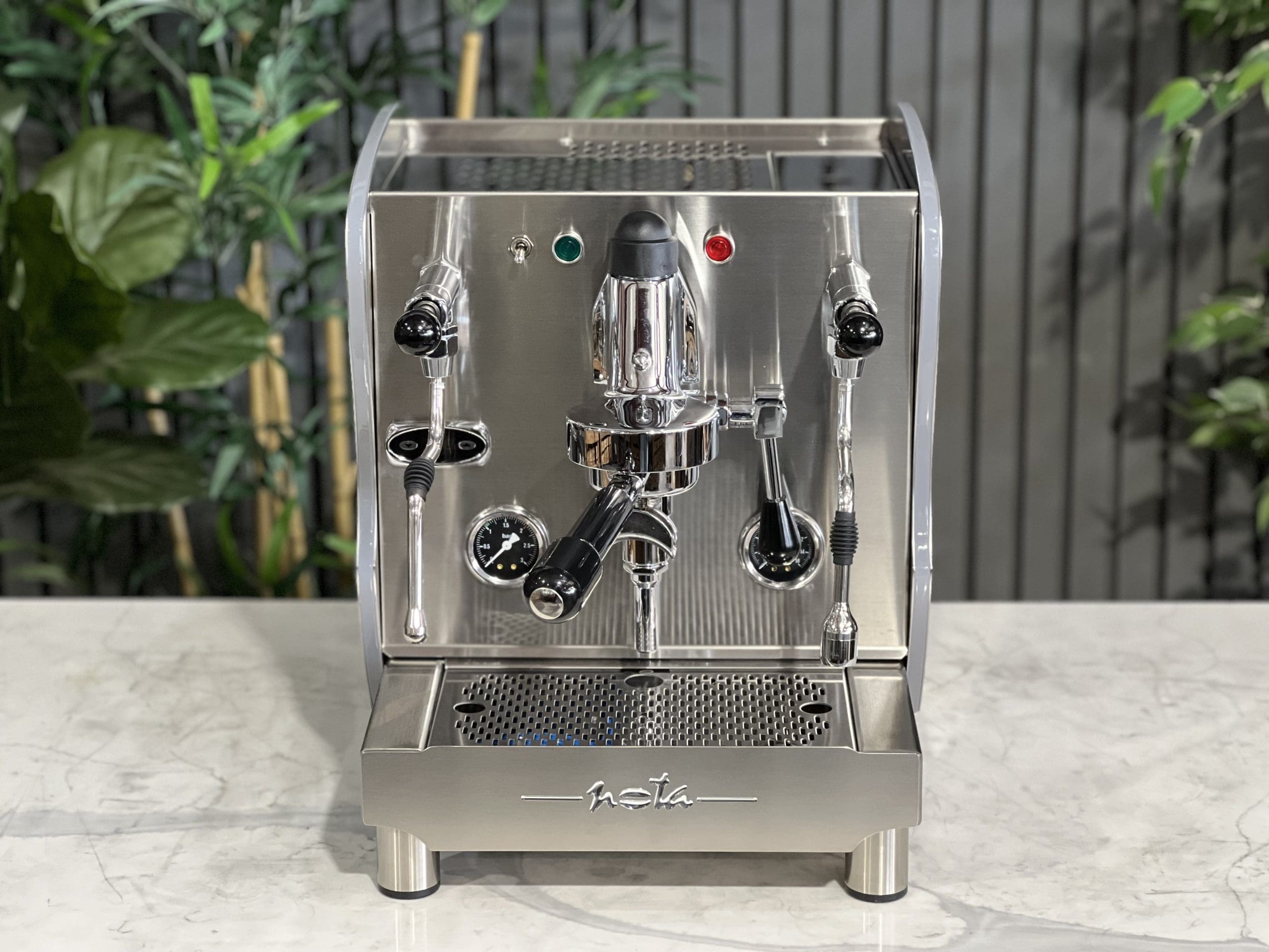 A brand new Orchestrale Nota PID 1 Group Coffee Machine with Stainless finish and Side Lights sitting on a marble bench. There is greenery in the background. The image is taken from the front of the machine so that you can see the front of the machine, control panel, buttons and group heads.