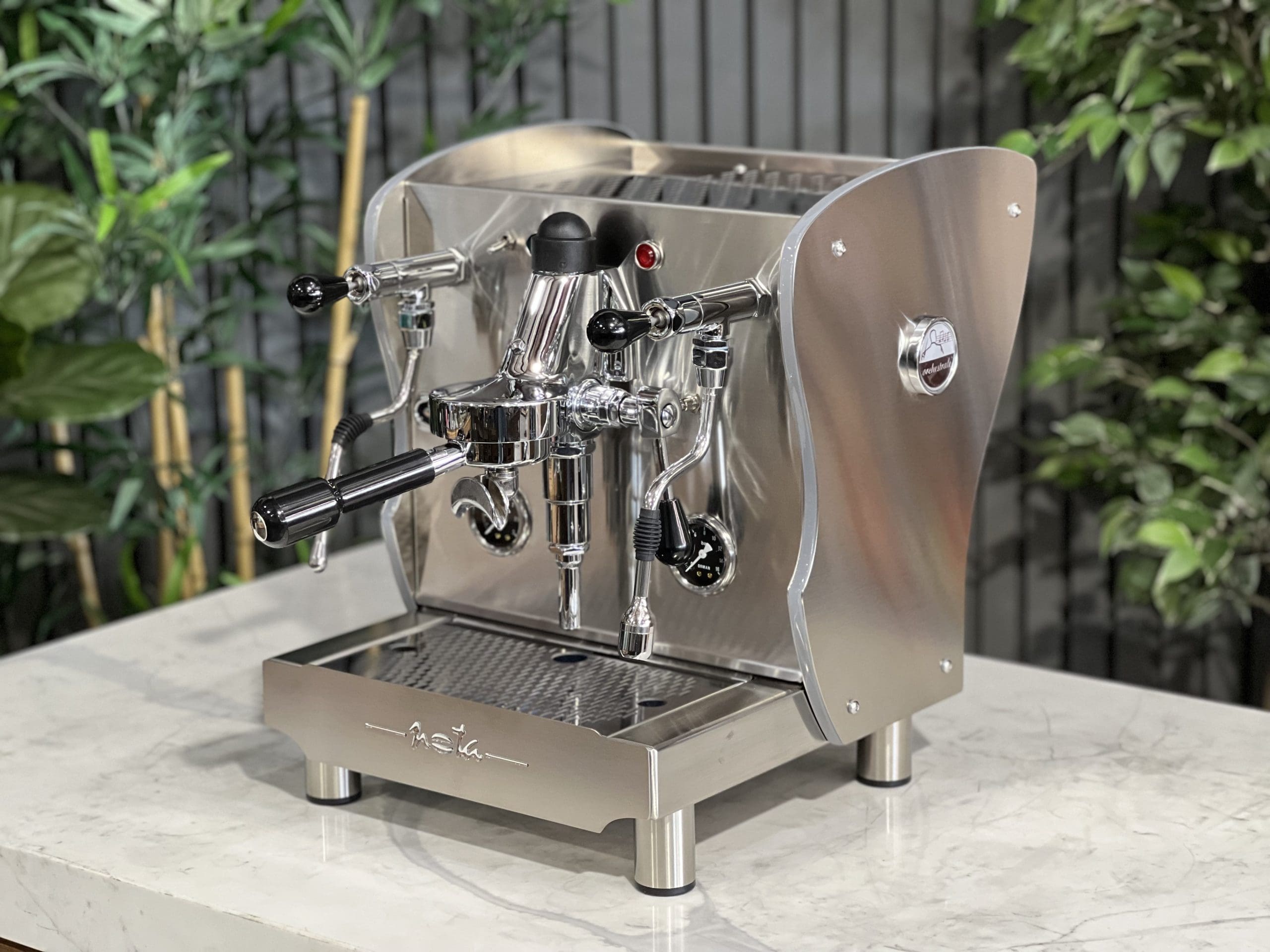 A brand new Orchestrale Nota PID 1 Group Coffee Machine with Stainless finish and Side Lights is sitting on a marble bench. There is greenery in the background. The image is taken from the front of the machine and the machine is sitting on an angle so that you can see the front of the machine and the side.