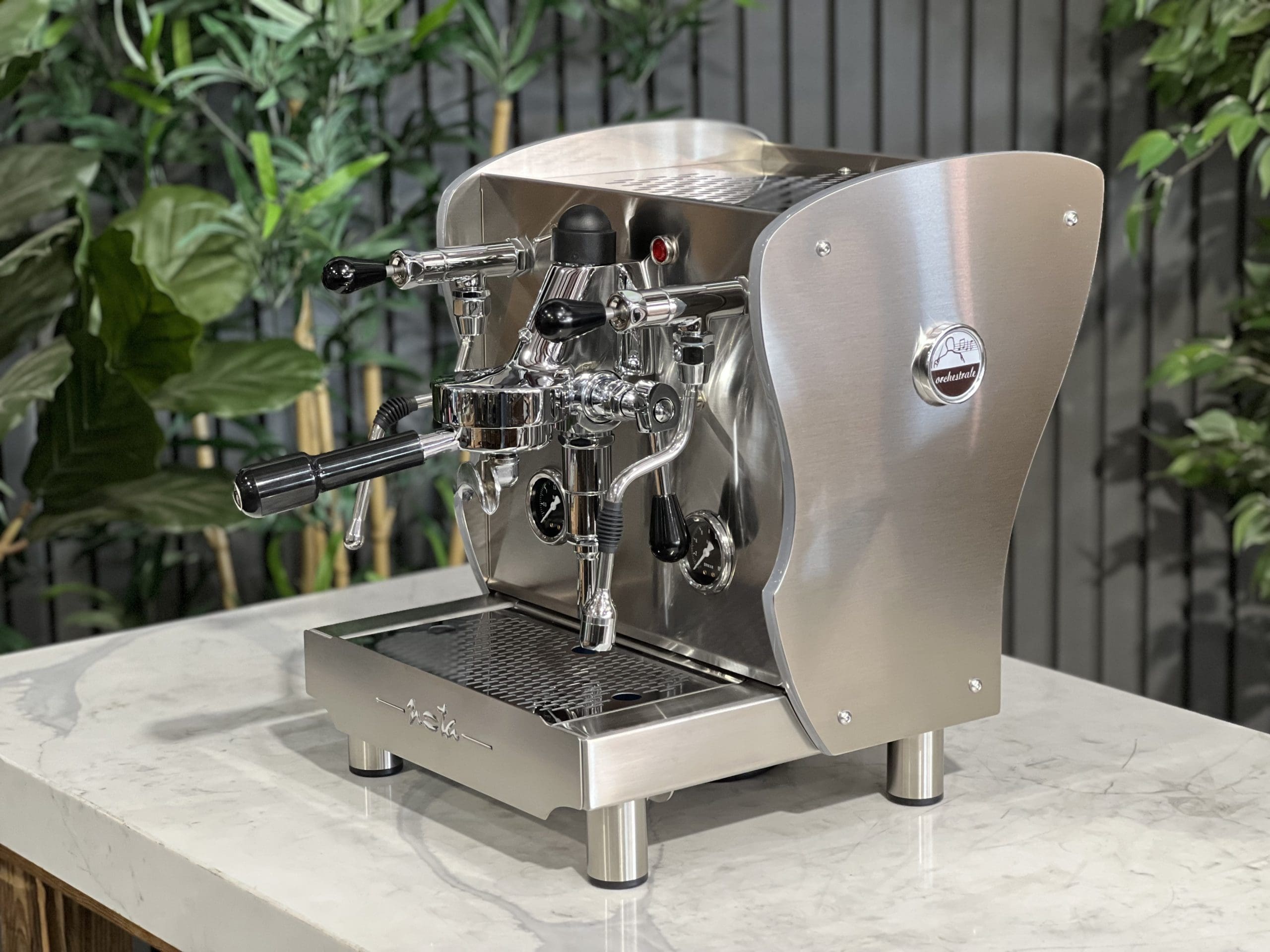 A brand new Orchestrale Nota PID 1 Group Coffee Machine with Stainless finish and Side Lights is sitting on a marble bench. There is greenery in the background. The image is taken from the front of the machine and the machine is sitting on an angle so that you can see the front of the machine and the side.