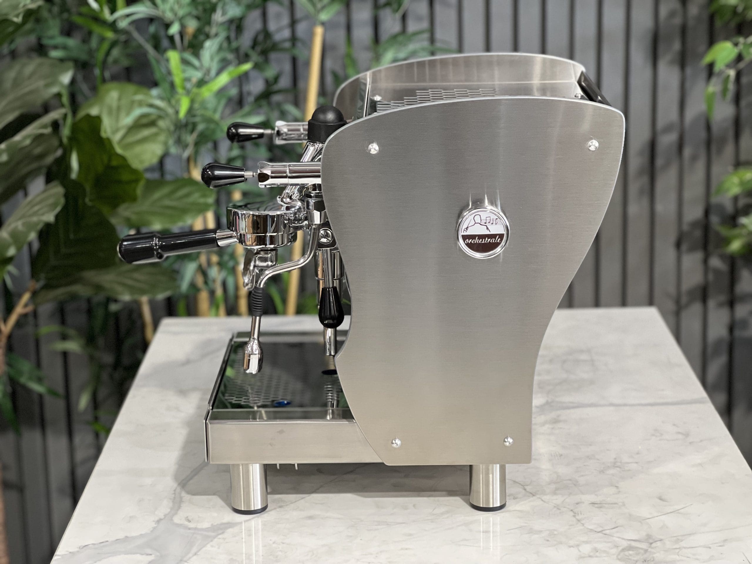 A brand new Orchestrale Nota PID 1 Group Coffee Machine with Stainless finish and Side Lights sitting on a marble bench. There is greenery in the background. The image is taken from the side of the machine.