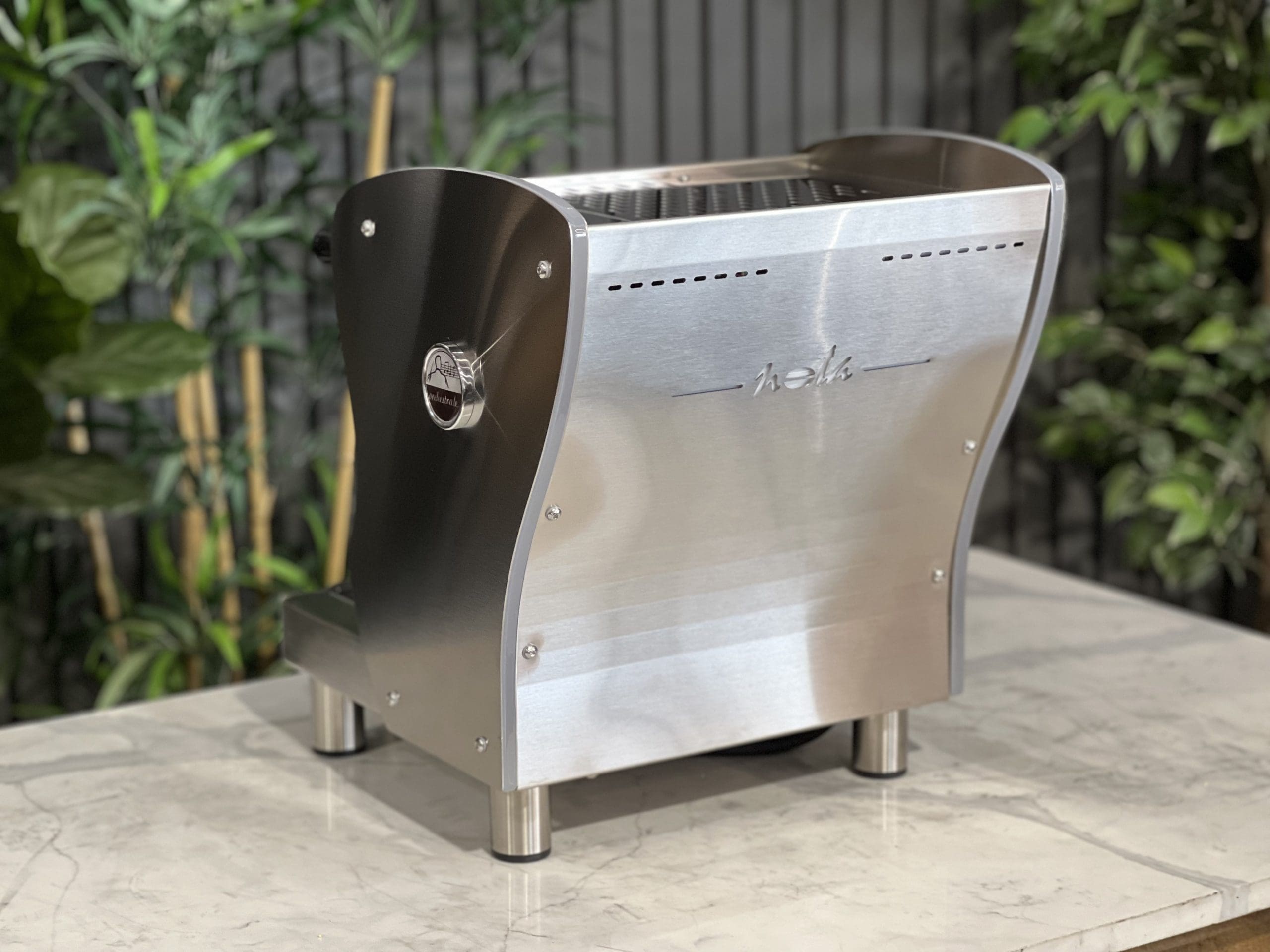 A brand new Orchestrale Nota PID 1 Group Coffee Machine with Stainless finish and Side Lights sitting on a marble bench. There is greenery in the background. The image is taken from behind the machine and the machine is sitting on an angle so that you can see the back of the machine and the side.