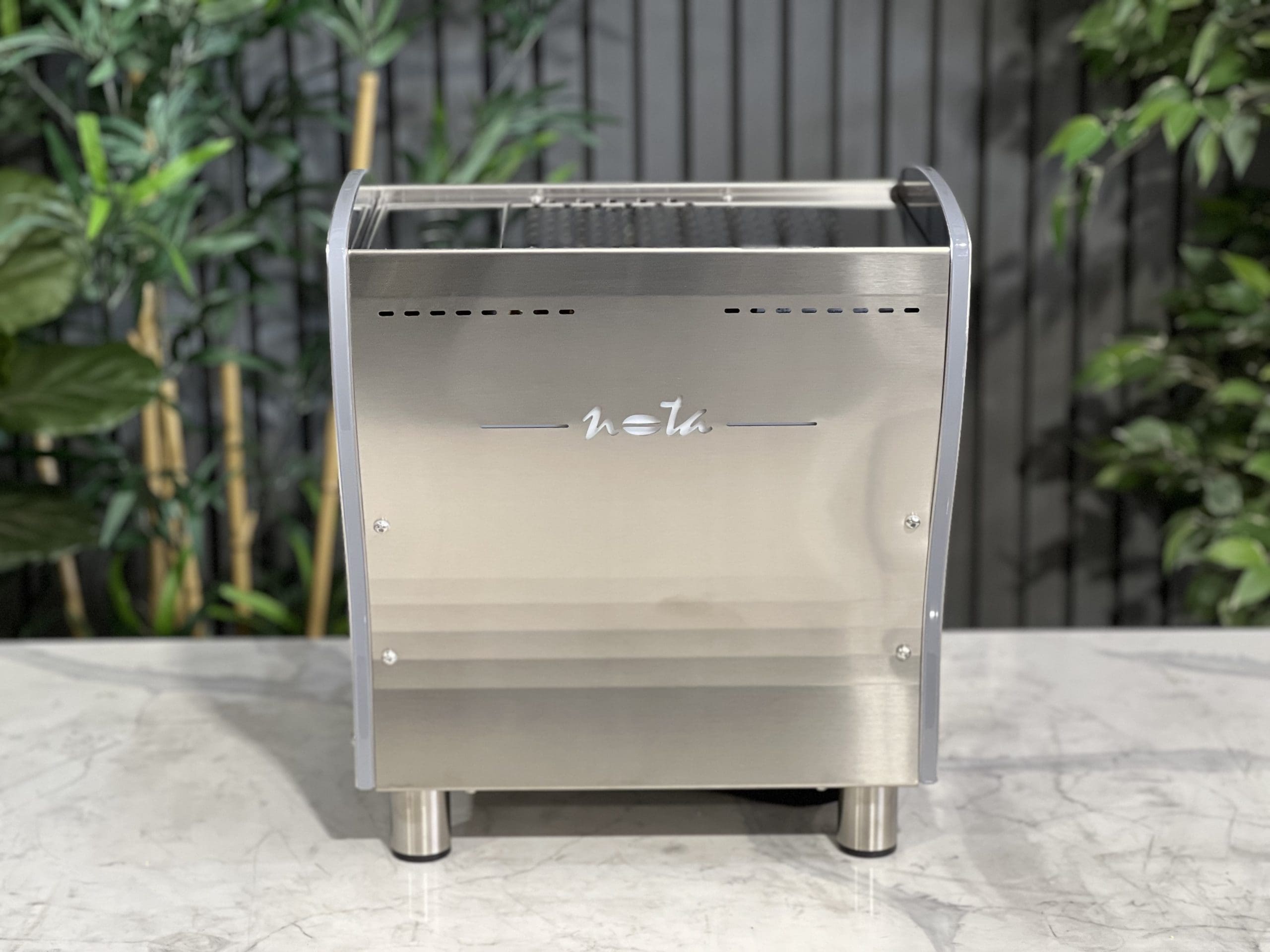 A brand new Orchestrale Nota PID 1 Group Coffee Machine with Stainless finish and Side Lights is sitting on a marble bench. There is greenery in the background. The image is taken from behind the machine so the back is visible.