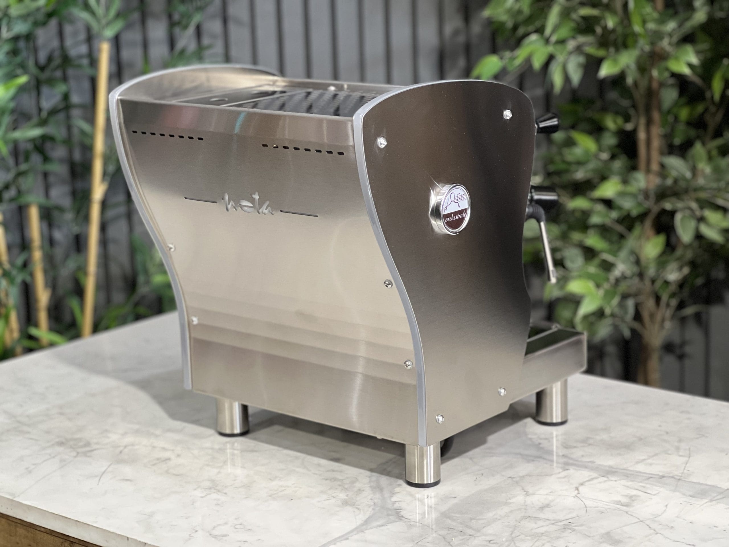 A brand new Orchestrale Nota PID 1 Group Coffee Machine with Stainless finish and Side Lights sitting on a marble bench. There is greenery in the background. The image is taken from behind the machine and the machine is sitting on an angle so that you can see the back of the machine and the side.