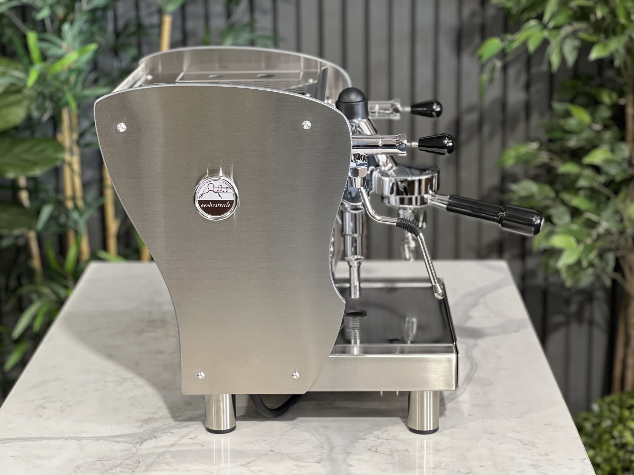 A brand new Orchestrale Nota PID 1 Group Coffee Machine with Stainless finish and Side Lights sitting on a marble bench. There is greenery in the background. The image is taken from the side of the machine.