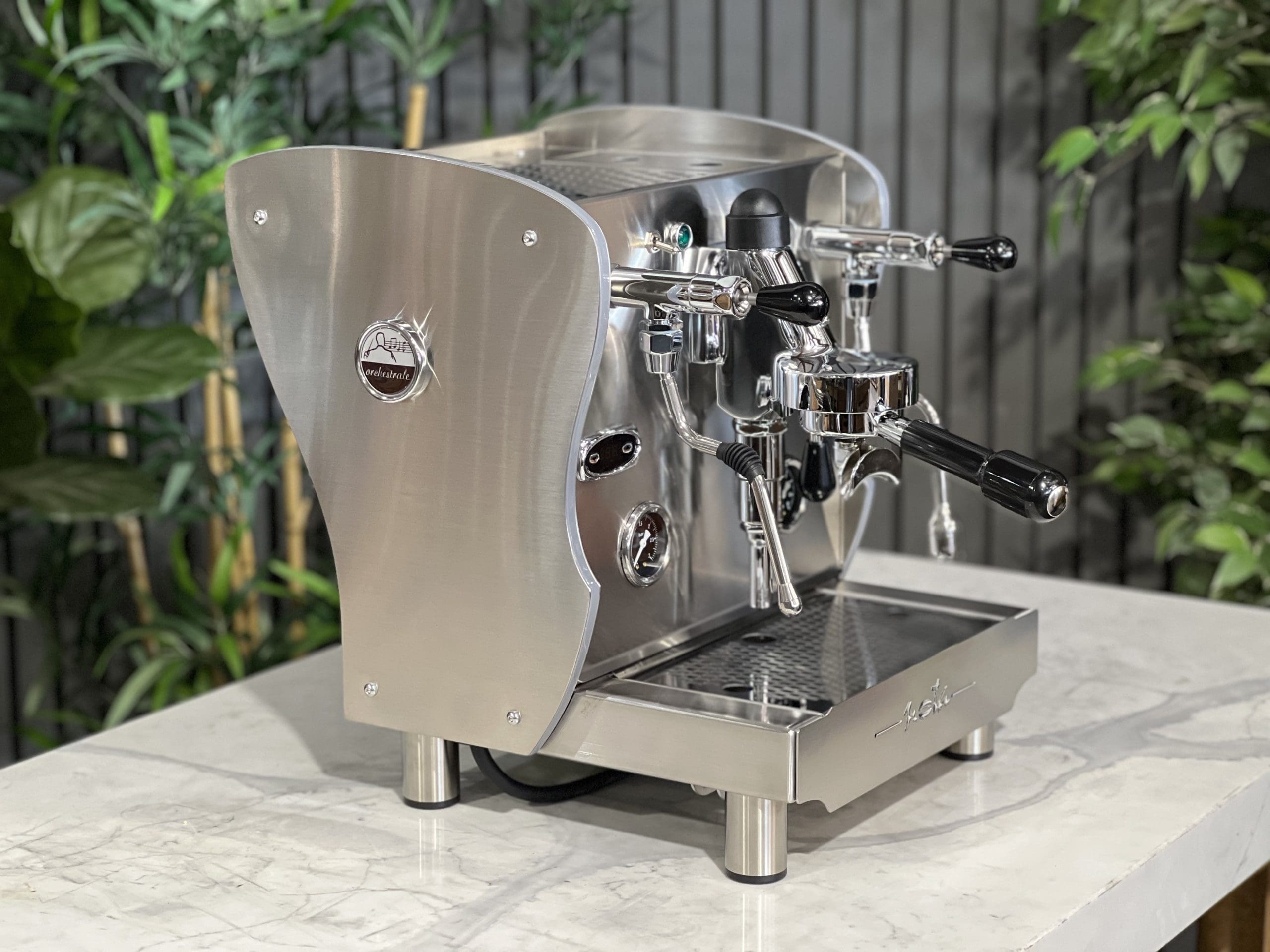 A brand new Orchestrale Nota PID 1 Group Coffee Machine with Stainless finish and Side Lights is sitting on a marble bench. There is greenery in the background. The image is taken from the front of the machine and the machine is sitting on an angle so that you can see the front of the machine and the side.