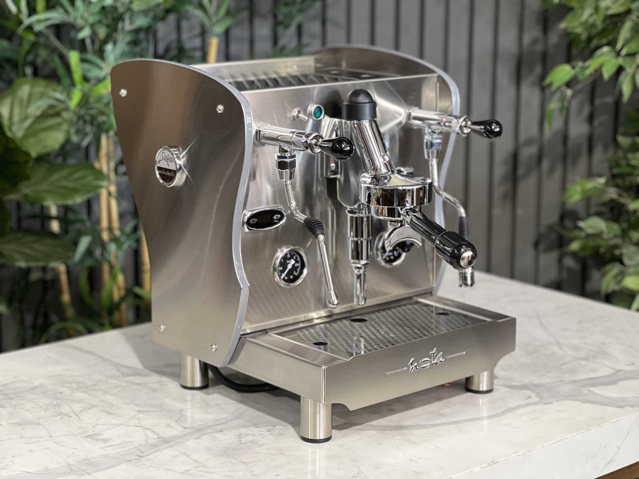 A brand new Orchestrale Nota PID 1 Group Coffee Machine with Stainless finish and Side Lights is sitting on a marble bench. There is greenery in the background. The image is taken from the front of the machine and the machine is sitting on an angle so that you can see the front of the machine and the side.