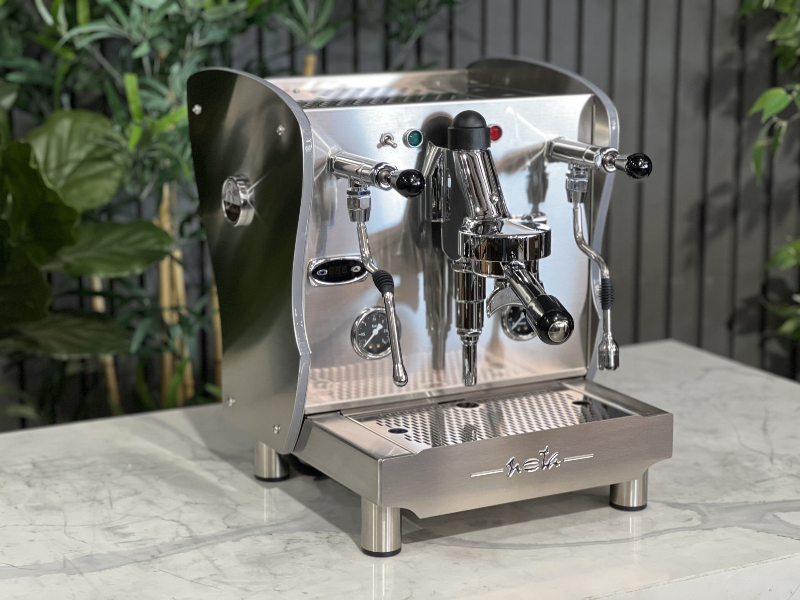 A brand new Orchestrale Nota PID 1 Group Coffee Machine with Stainless finish and Side Lights is sitting on a marble bench. There is greenery in the background. The image is taken from the front of the machine and the machine is sitting on an angle so that you can see the front of the machine and the side.
