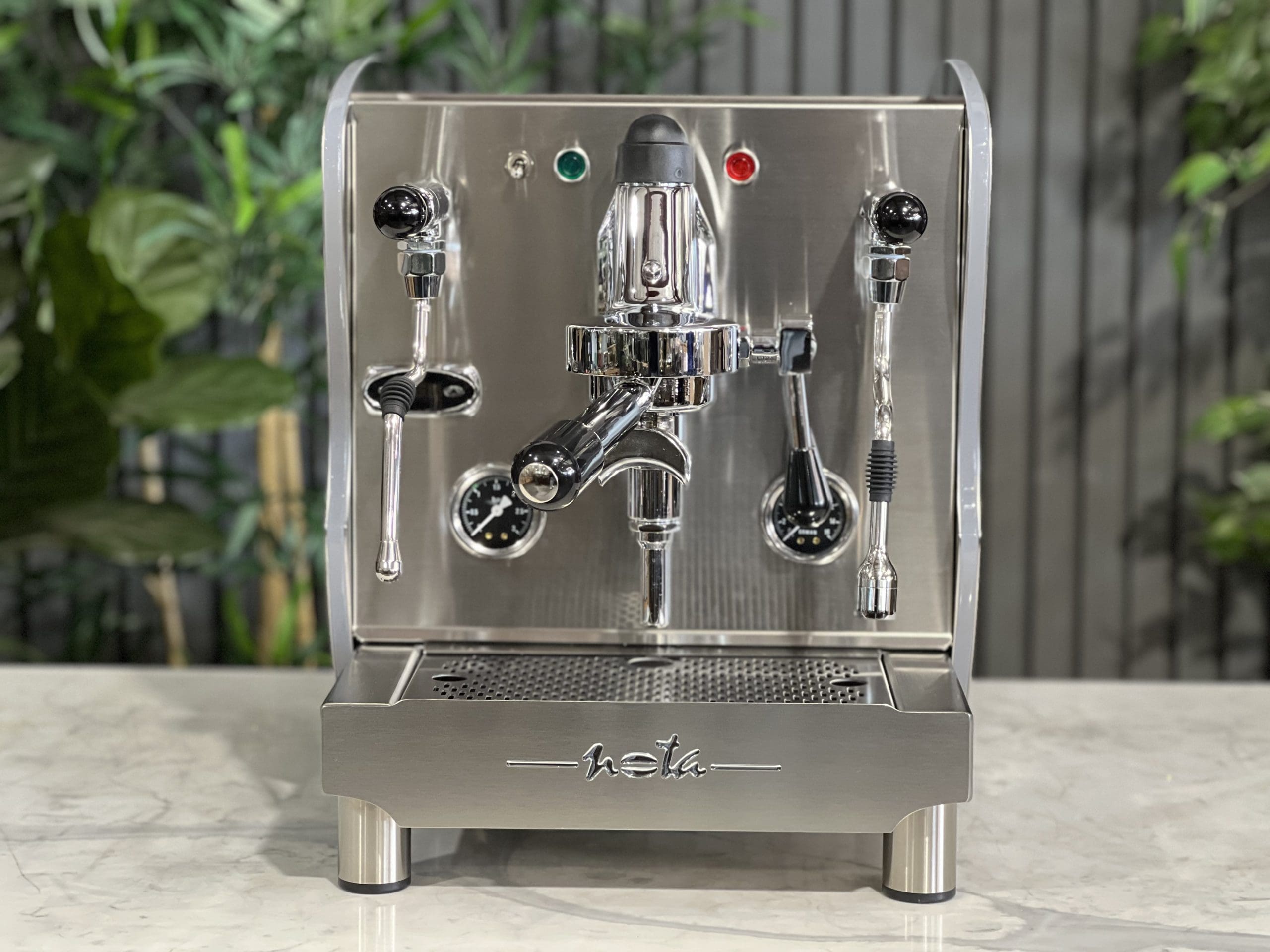 A brand new Orchestrale Nota PID 1 Group Coffee Machine with Stainless finish and Side Lights sitting on a marble bench. There is greenery in the background. The image is taken from the front of the machine so that you can see the front of the machine, control panel, buttons and group heads.