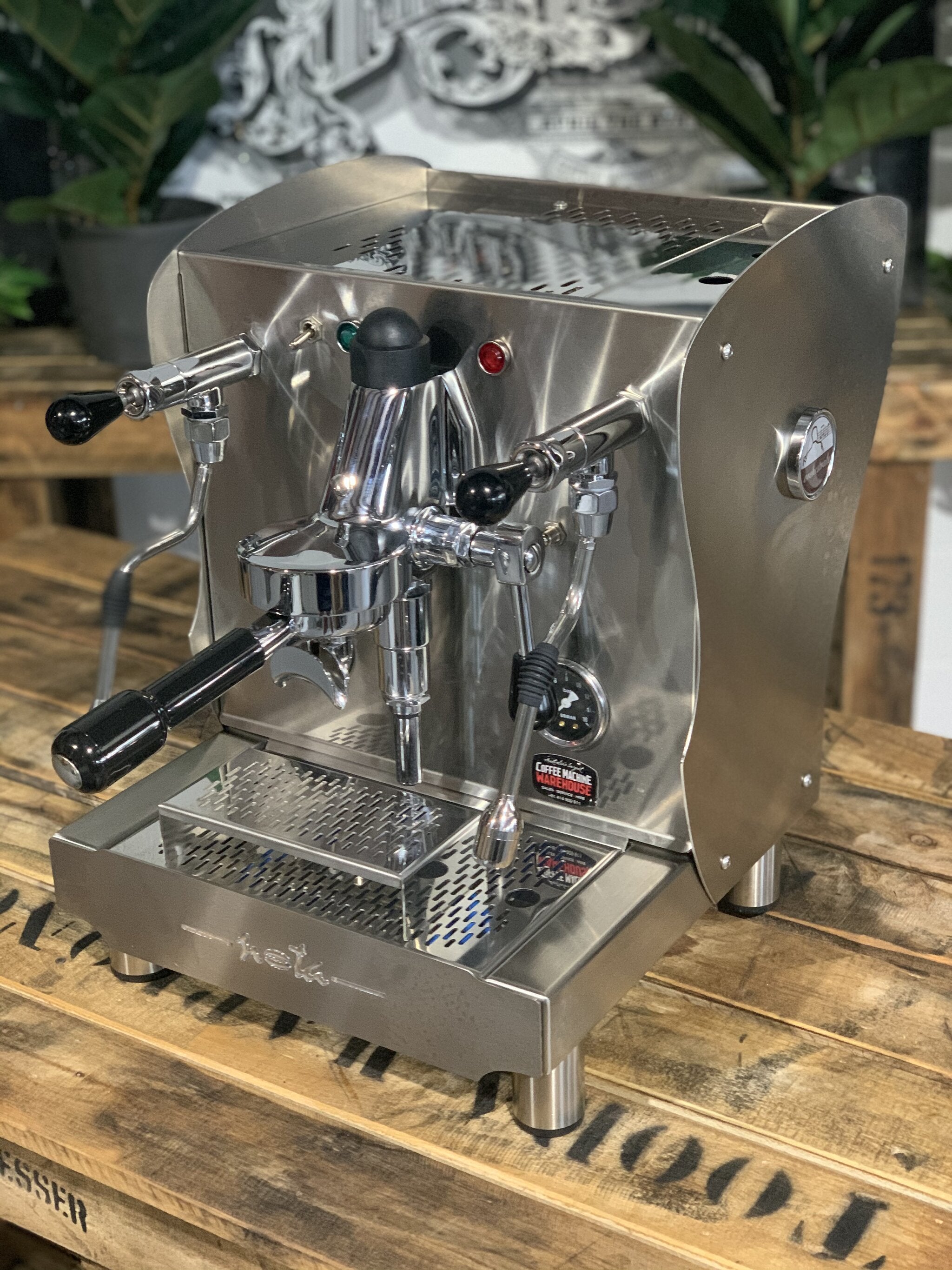 A brand new Orchestrale Nota 1 Group Espresso Coffee Machine with Stainless finish is sitting on a wooden bench. There is greenery in the background. The image is taken from the front of the machine and the machine is sitting on an angle so that you can see the front of the machine and the side.
