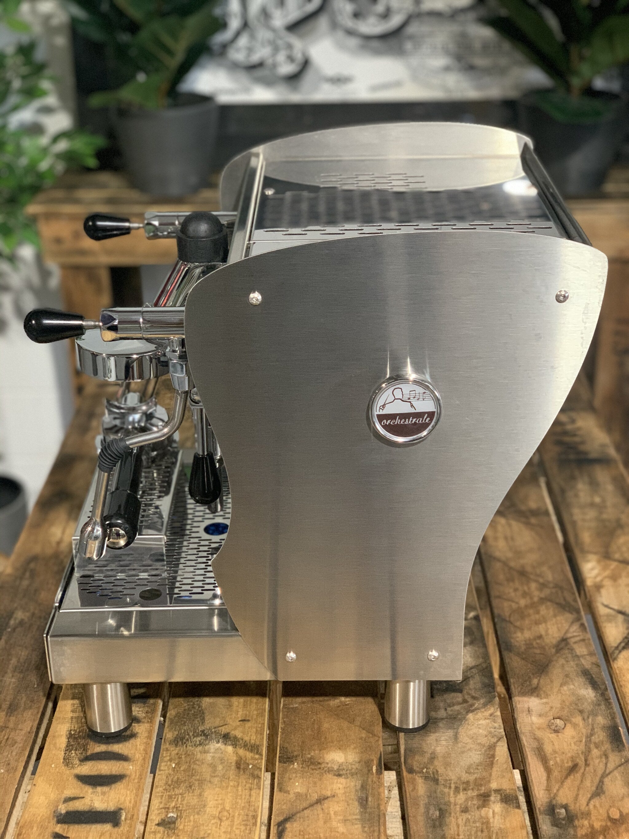 A brand new Orchestrale Nota 1 Group Espresso Coffee Machine with Stainless finish sitting on a wooden bench. There is greenery in the background. The image is taken from the side of the machine.