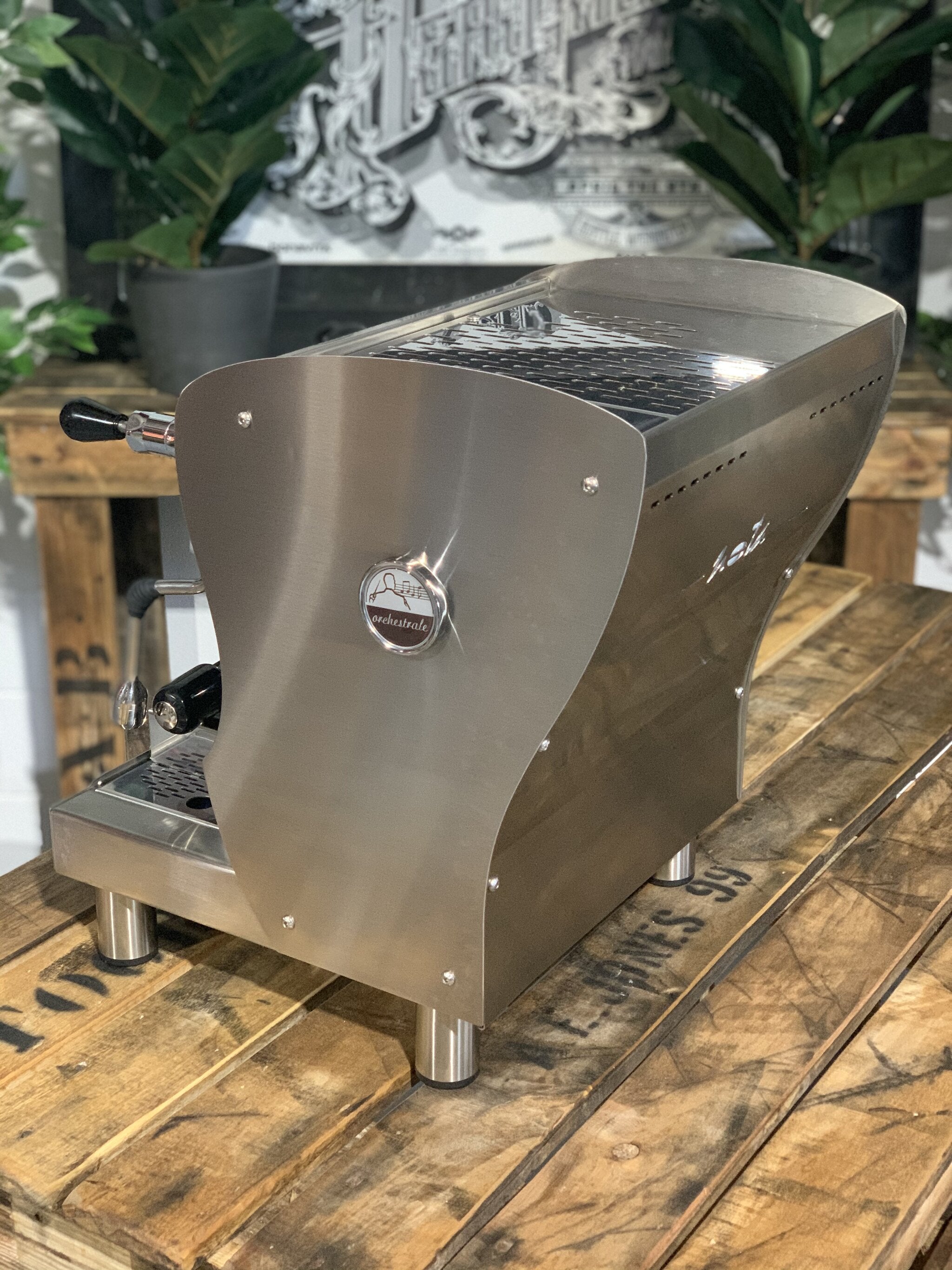 A brand new Orchestrale Nota 1 Group Espresso Coffee Machine with Stainless finishsitting on a wooden bench. There is greenery in the background. The image is taken from behind the machine and the machine is sitting on an angle so that you can see the back of the machine and the side.