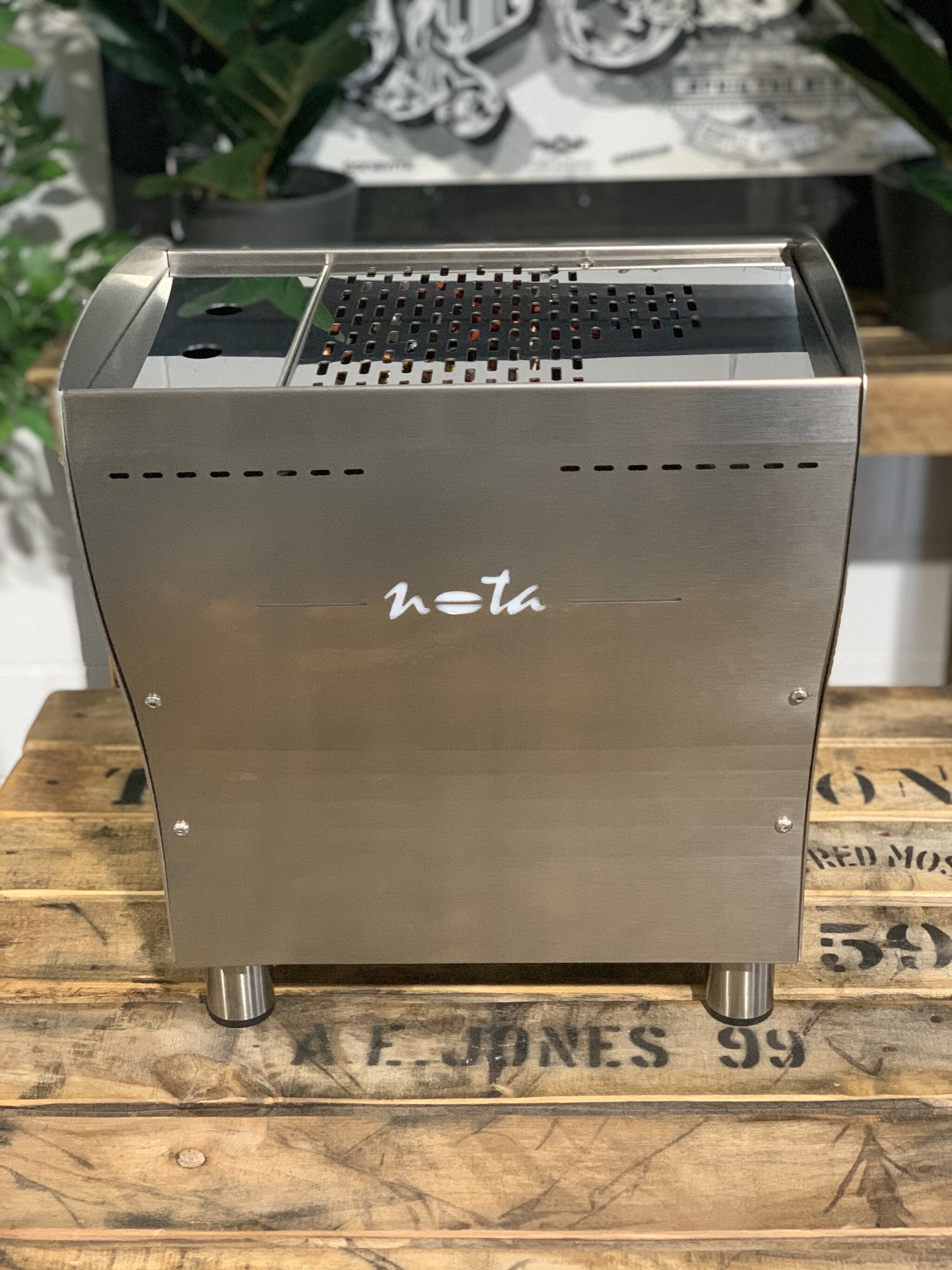 A brand new Orchestrale Nota 1 Group Espresso Coffee Machine with Stainless finish s sitting on a wooden bench. There is greenery in the background. The image is taken from behind the machine so the back is visible. The back of the machine is a stainless steel panel with the Nota logo.