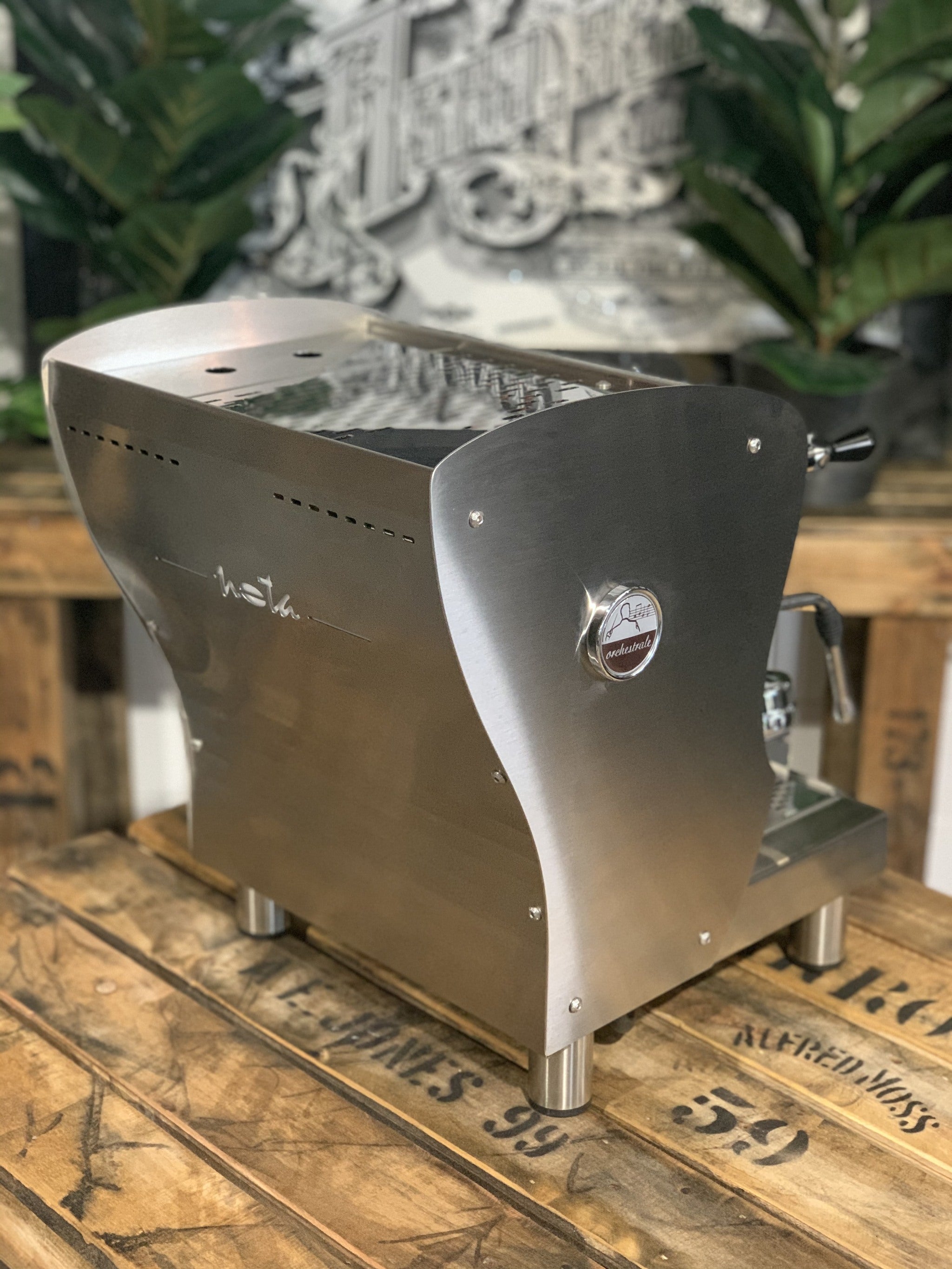 A brand new Orchestrale Nota 1 Group Espresso Coffee Machine with Stainless finish sitting on a wooden bench. There is greenery in the background. The image is taken from behind the machine and the machine is sitting on an angle so that you can see the back of the machine and the side.