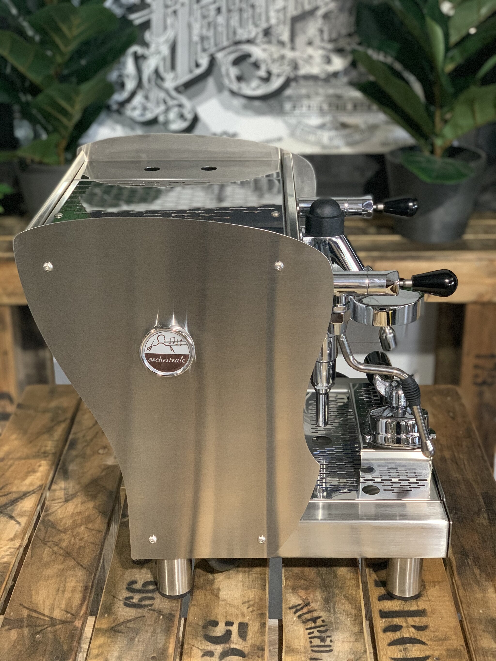 A brand new Orchestrale Nota 1 Group Espresso Coffee Machine with Stainless finish sitting on a wooden bench. There is greenery in the background. The image is taken from the side of the machine.