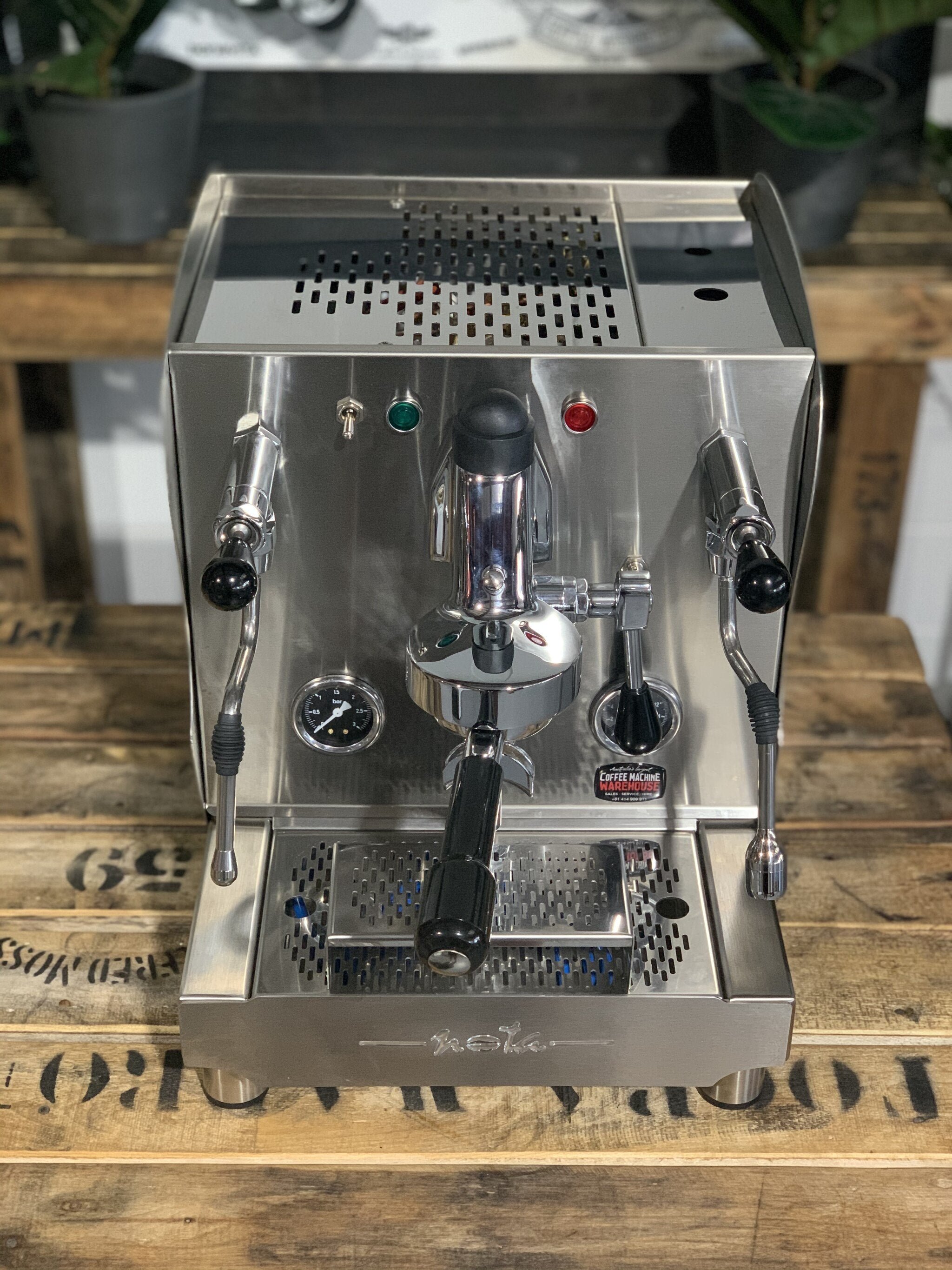 A brand new Orchestrale Nota 1 Group Espresso Coffee Machine with Stainless finish sitting on a wooden bench. There is greenery in the background. The image is taken from the front of the machine but a higher angle so that you can see the front of the machine and also the top of the machine.