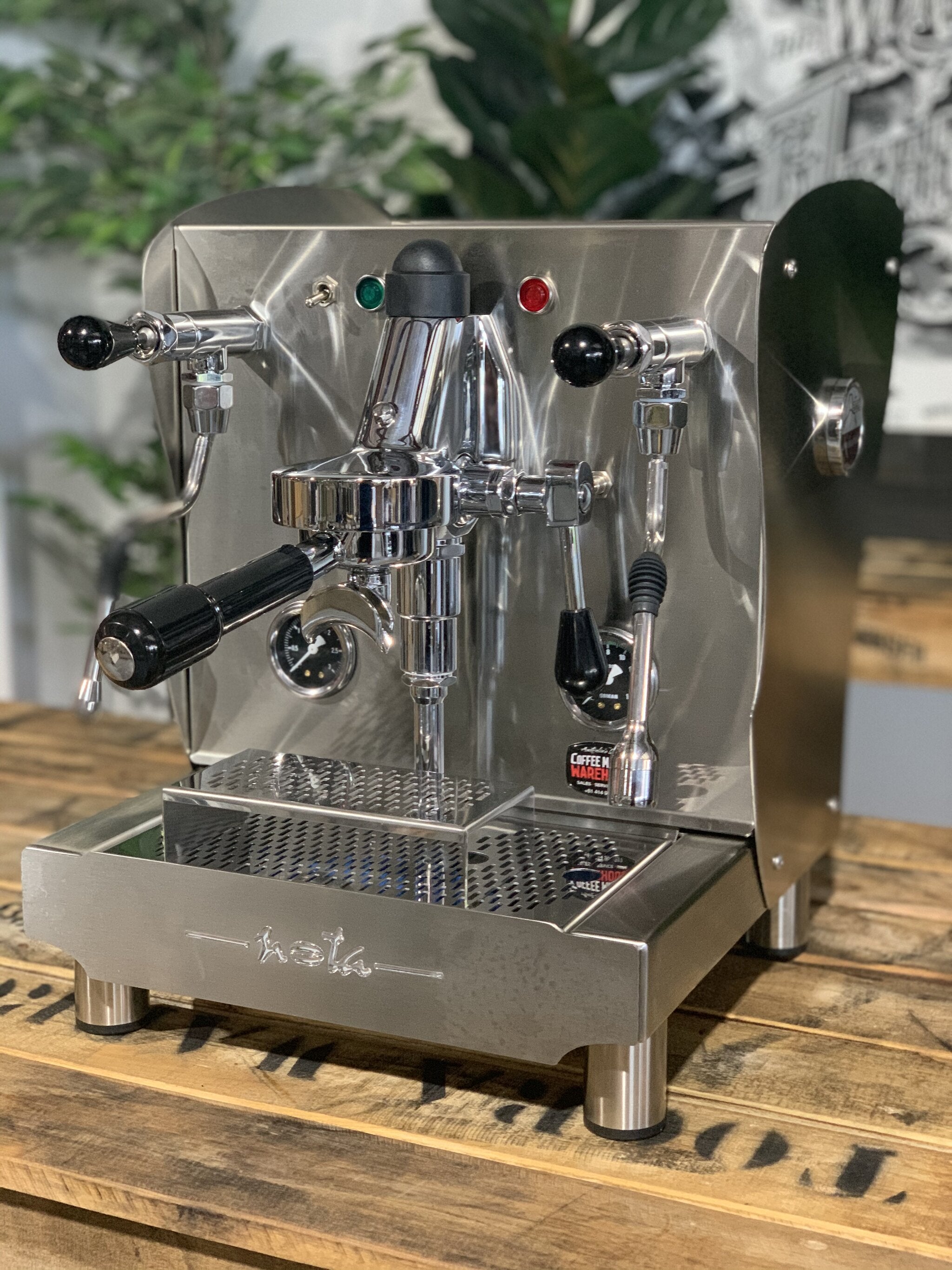 A brand new Orchestrale Nota 1 Group Espresso Coffee Machine with Stainless finish is sitting on a wooden bench. There is greenery in the background. The image is taken from the front of the machine and the machine is sitting on an angle so that you can see the front of the machine and the side.