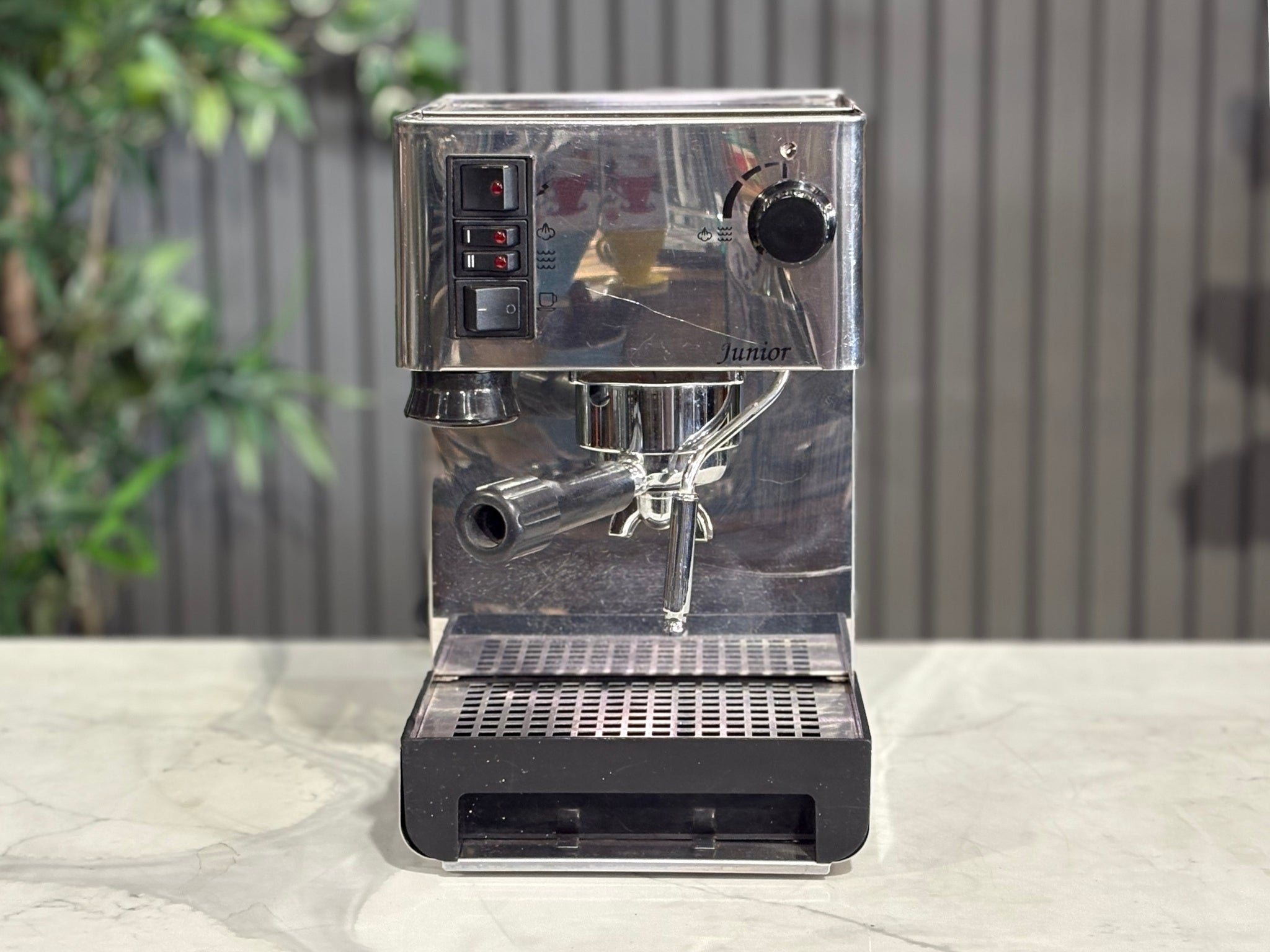 A Quaha Lelit Junior Serie II 1 Group Espresso Coffee Machine with Stainless finish  sitting on a marble bench. There is greenery in the background. The image is taken from the front of the machine so that you can see the front of the machine, control panel, buttons and group heads.