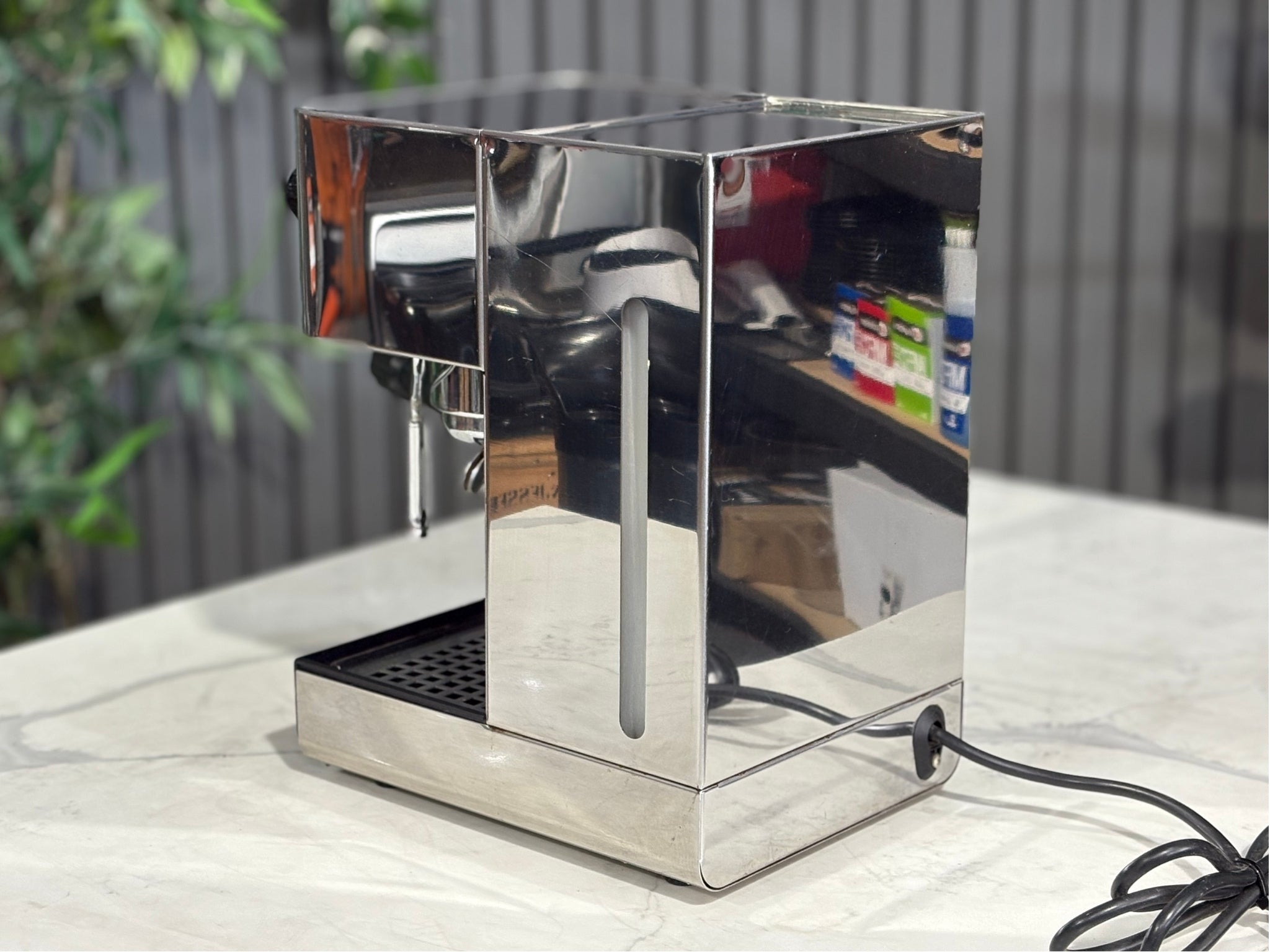 A Quaha Lelit Junior Serie II 1 Group Espresso Coffee Machine with Stainless finish sitting on a marble bench. There is greenery in the background. The image is taken from behind the machine and the machine is sitting on an angle so that you can see the back of the machine and the side.