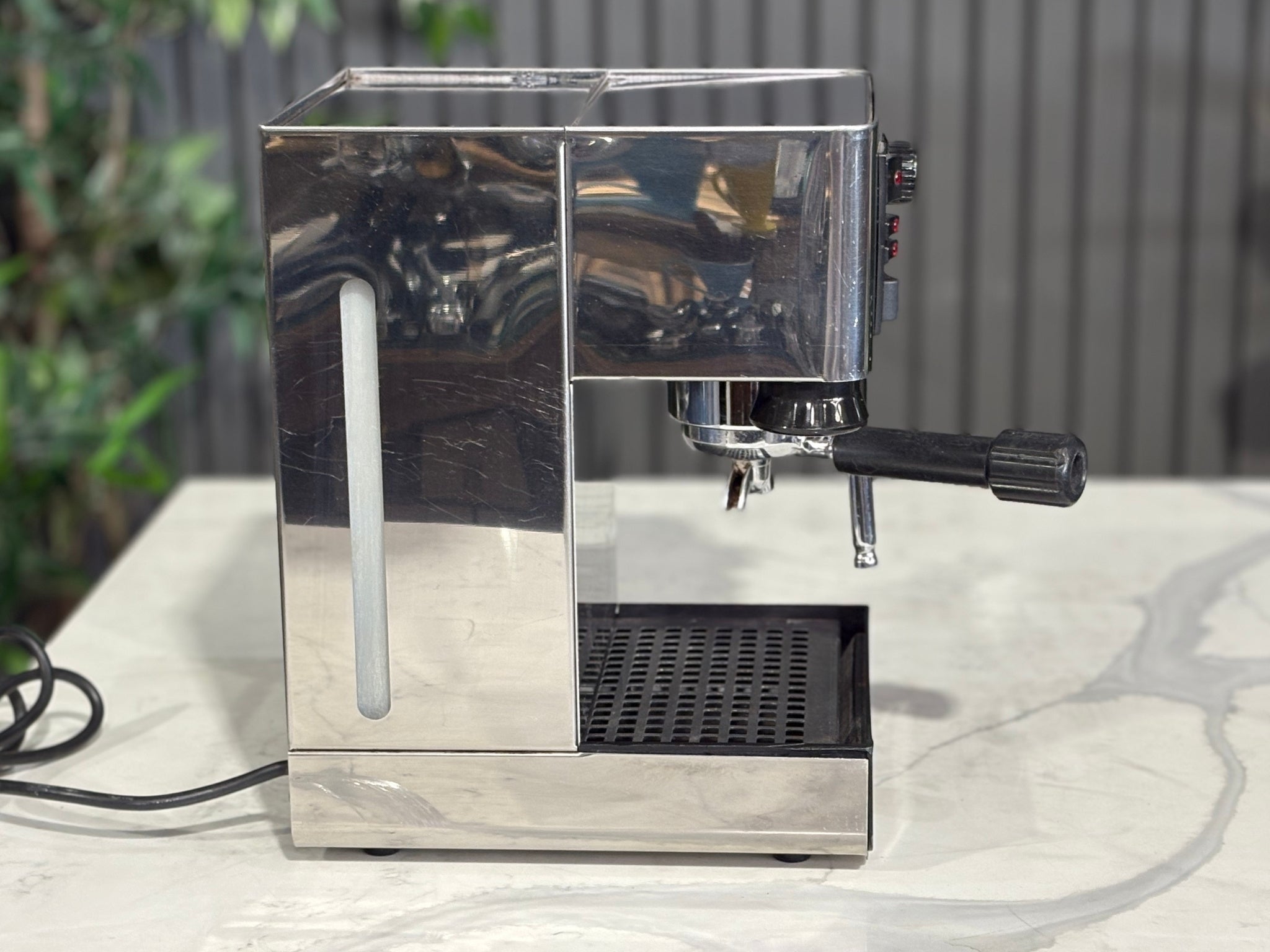 A Quaha Lelit Junior Serie II 1 Group Espresso Coffee Machine with Stainless finish  sitting on a marble bench. There is greenery in the background. The image is taken from the side of the machine.