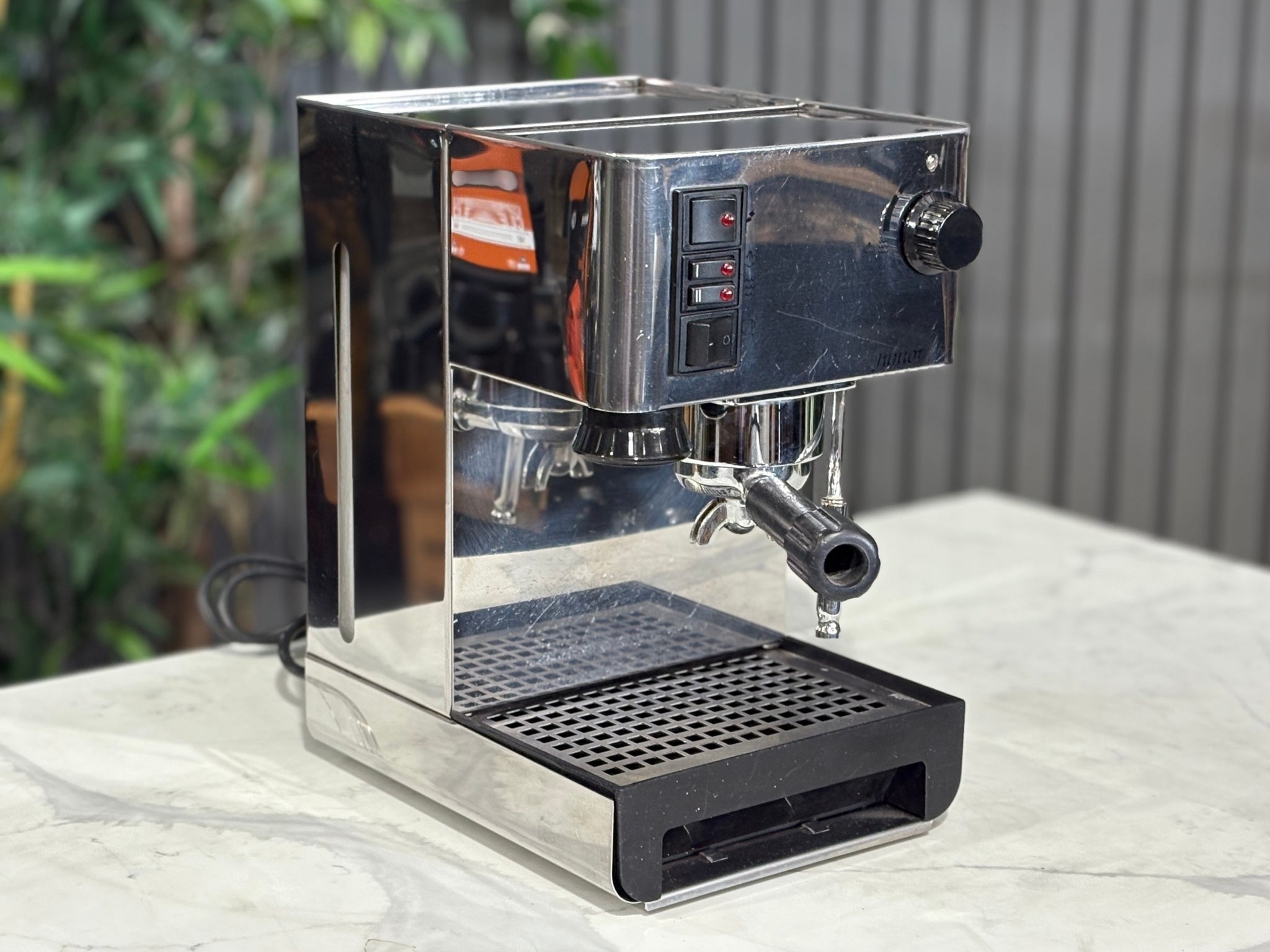 A Quaha Lelit Junior Serie II 1 Group Espresso Coffee Machine with Stainless finish is sitting on a marble bench. There is greenery in the background. The image is taken from the front of the machine and the machine is sitting on an angle so that you can see the front of the machine and the side.