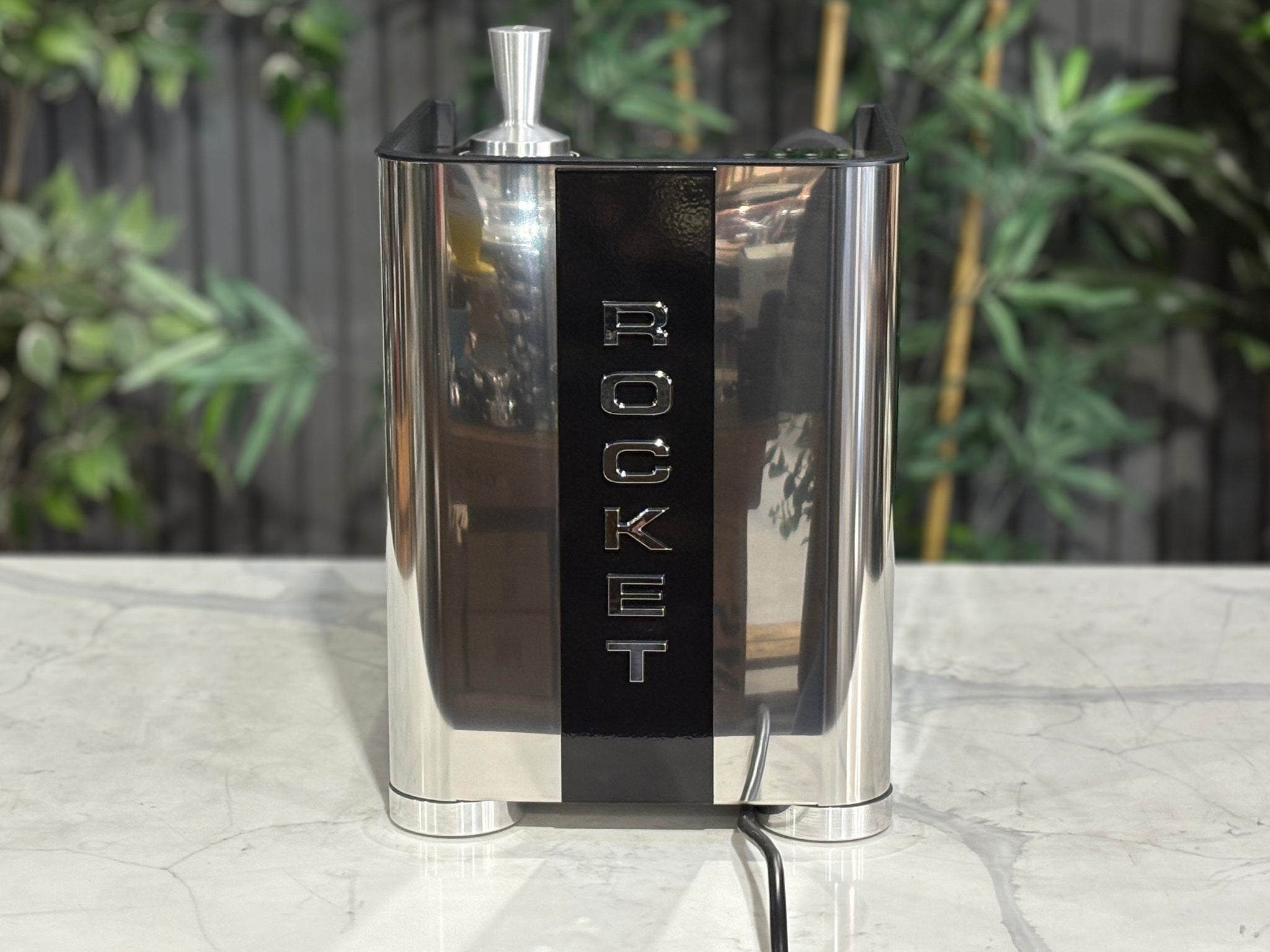 Rocket Appartamento TCA 1 Group Coffee Machine Stainless with Black Cutouts - New, rear view showing the vertical Rocket logo, rounded edges, and black cutouts on the stainless steel body on a marble bench with greenery in the background.