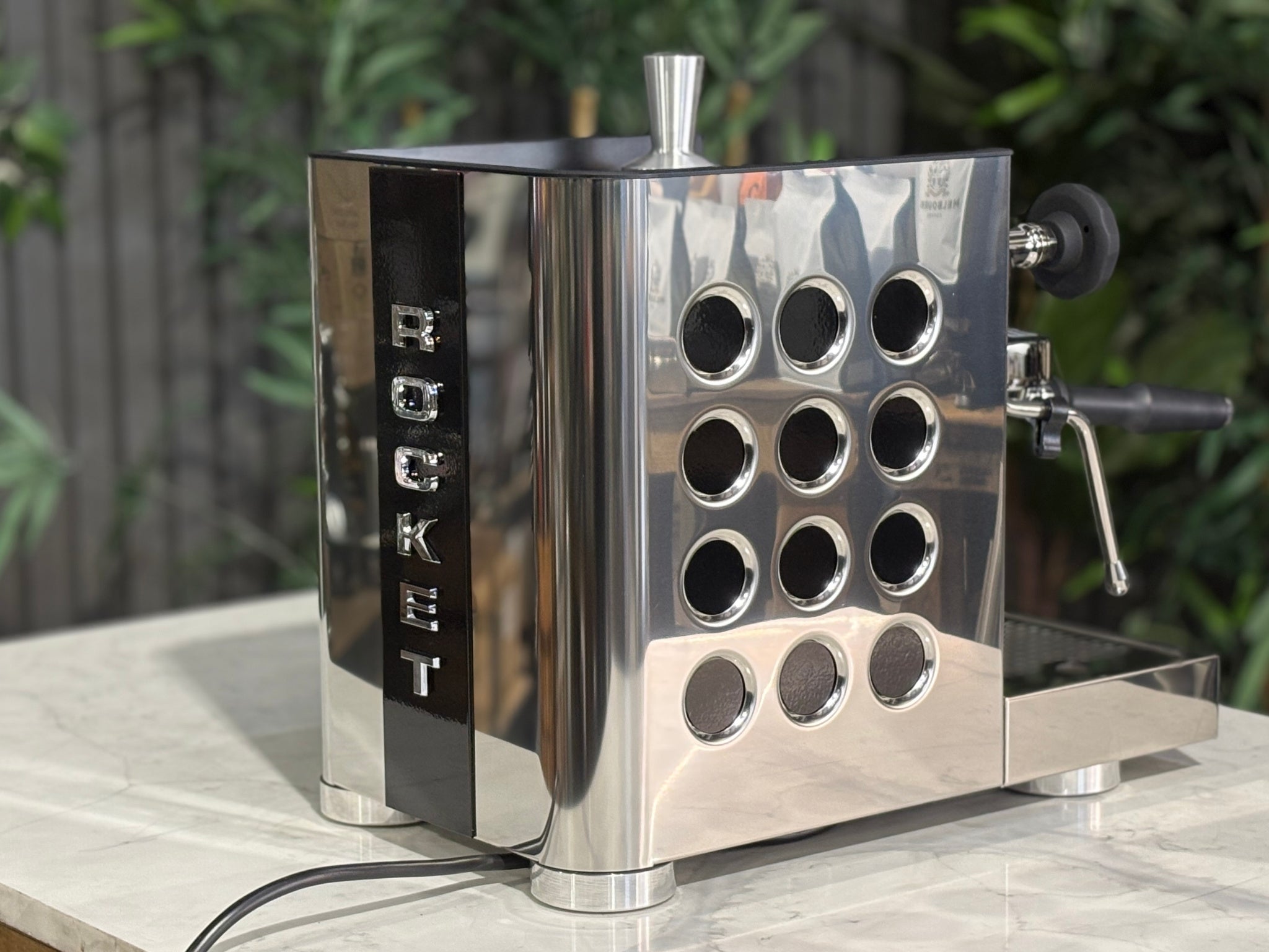 Rocket Appartamento TCA 1 Group Coffee Machine Stainless with Black Cutouts - New, angled rear view showing the vertical Rocket logo, black cutouts, and rounded stainless steel edges on a marble bench with greenery in the background.
