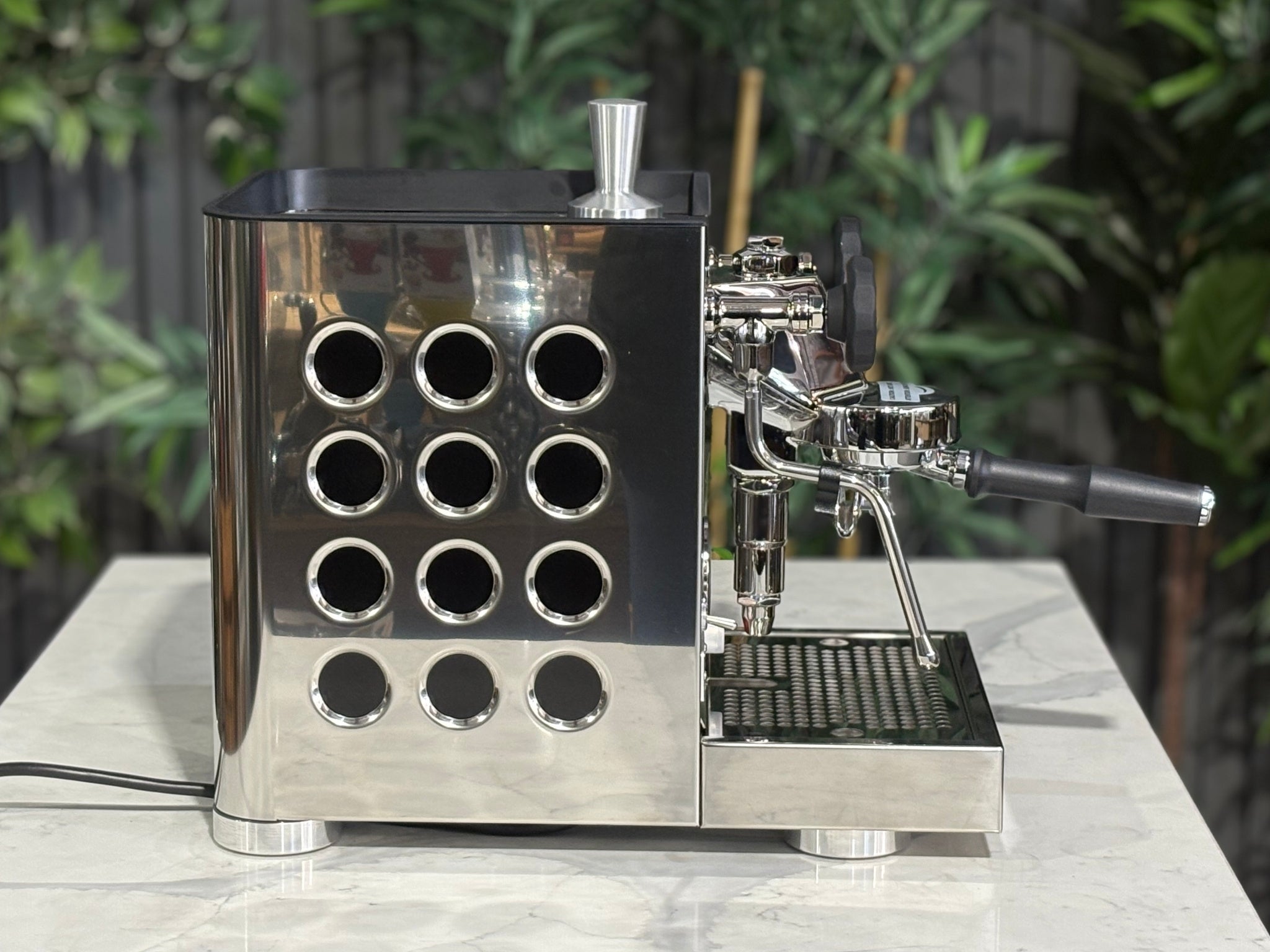 Rocket Appartamento TCA 1 Group Coffee Machine Stainless with Black Cutouts - New, side view displaying the circular debossed panel, rounded edges, and stainless steel body with black cutouts on a marble bench with greenery in the background.
