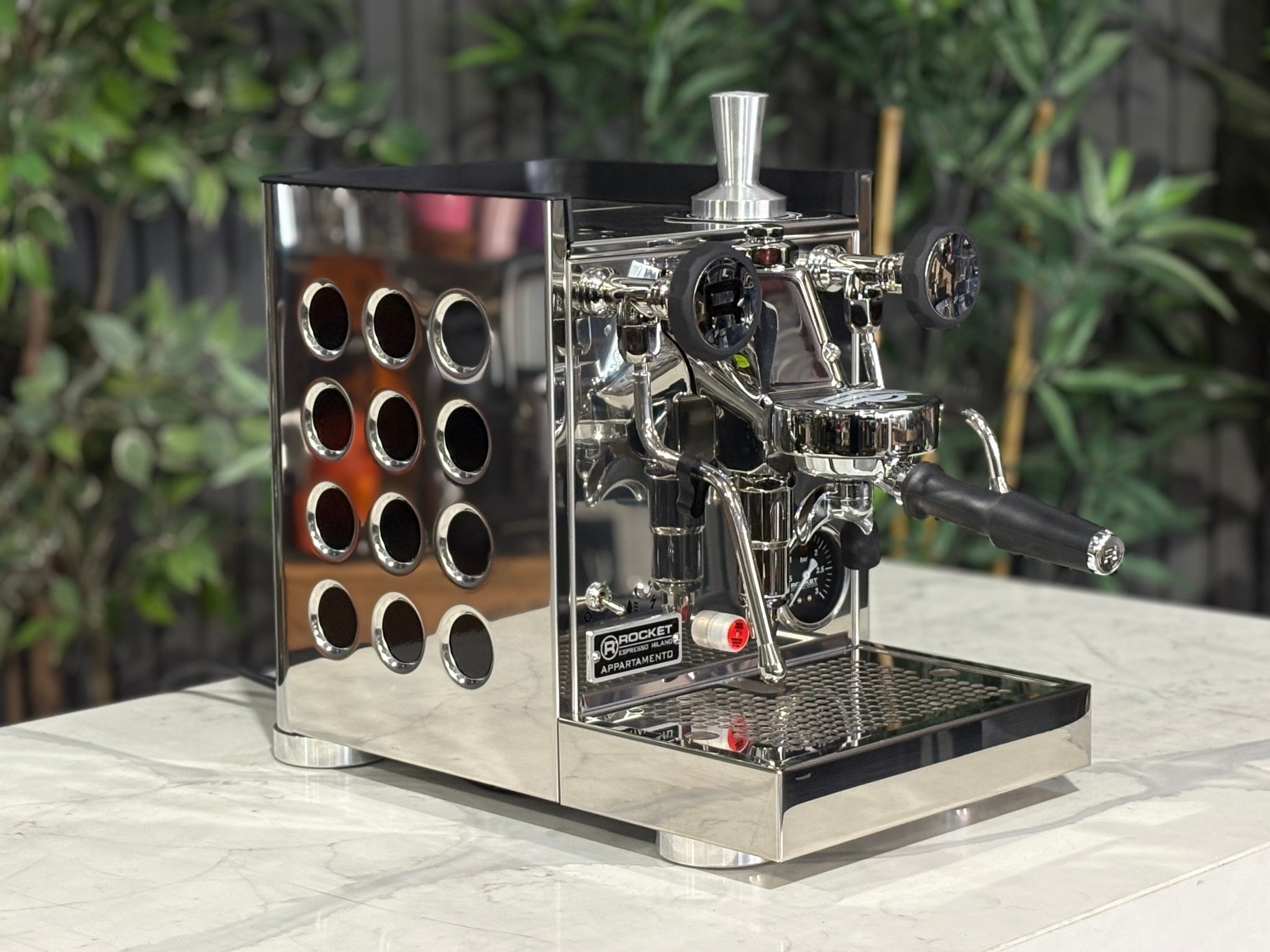 Rocket Appartamento TCA 1 Group Coffee Machine Stainless with Black Cutouts - New, angled view showing the E61 group head, high cup clearance, drip tray, and stainless steel body with black cutouts on a marble bench with greenery in the background.