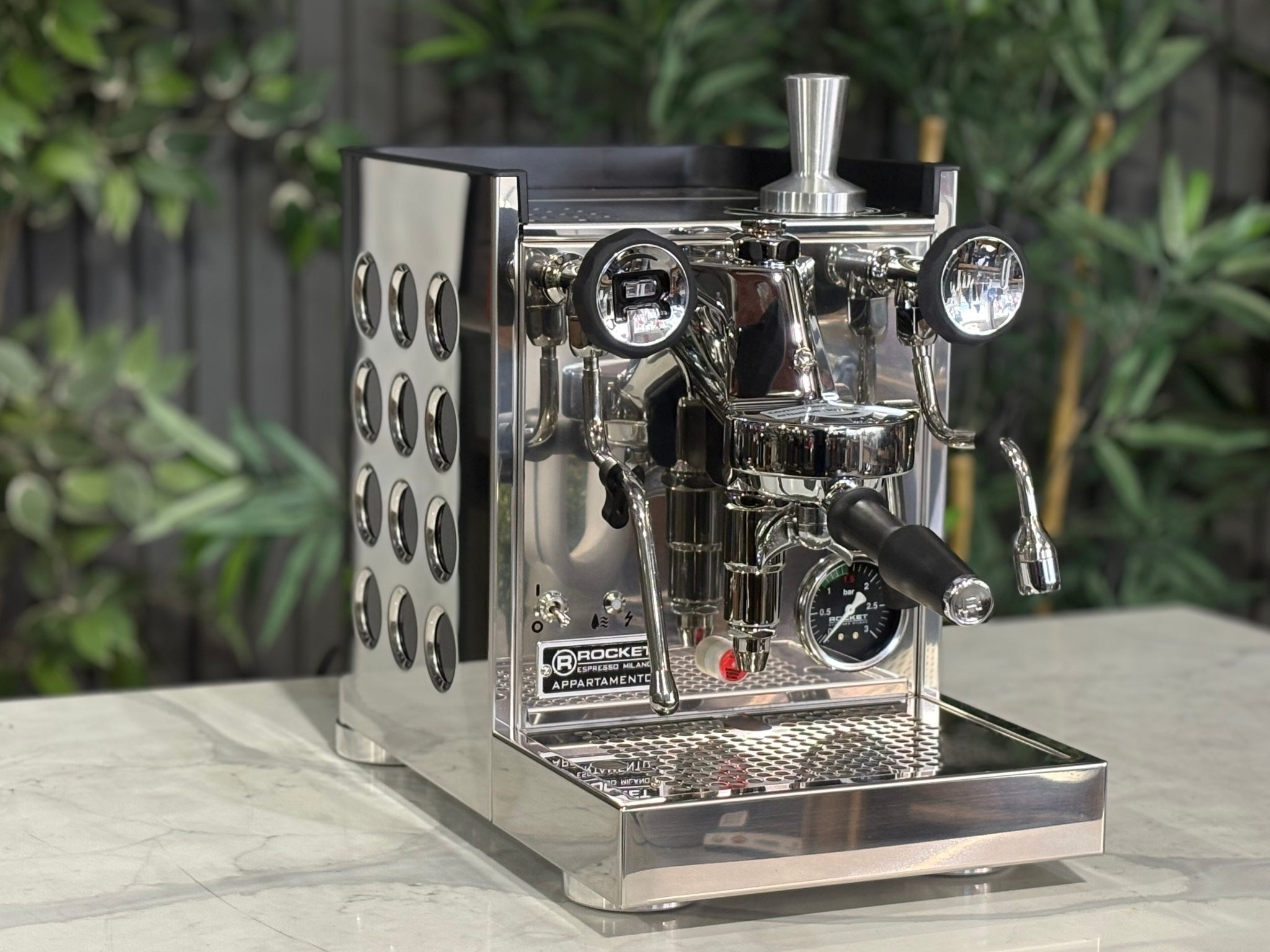 Rocket Appartamento TCA 1 Group Coffee Machine Stainless with Black Cutouts - New, angled view showing the E61 group head, high cup clearance, drip tray, and stainless steel body with black cutouts on a marble bench with greenery in the background.