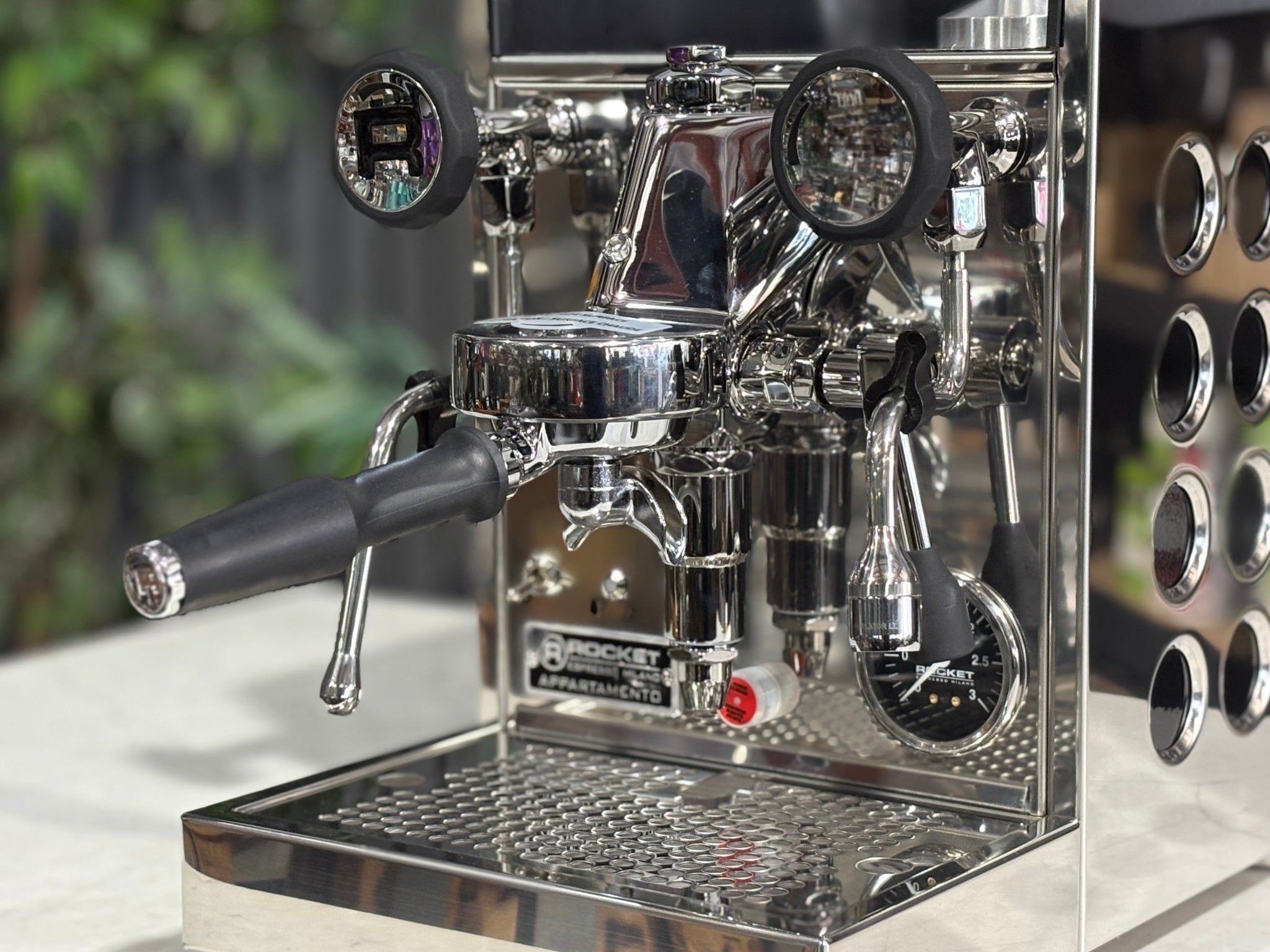 Rocket Appartamento TCA 1 Group Coffee Machine Stainless with Black Cutouts - New, close-up angled view showing the E61 group head, control lever, drip tray, and stainless steel finish with black cutouts on a marble bench with greenery in the background.