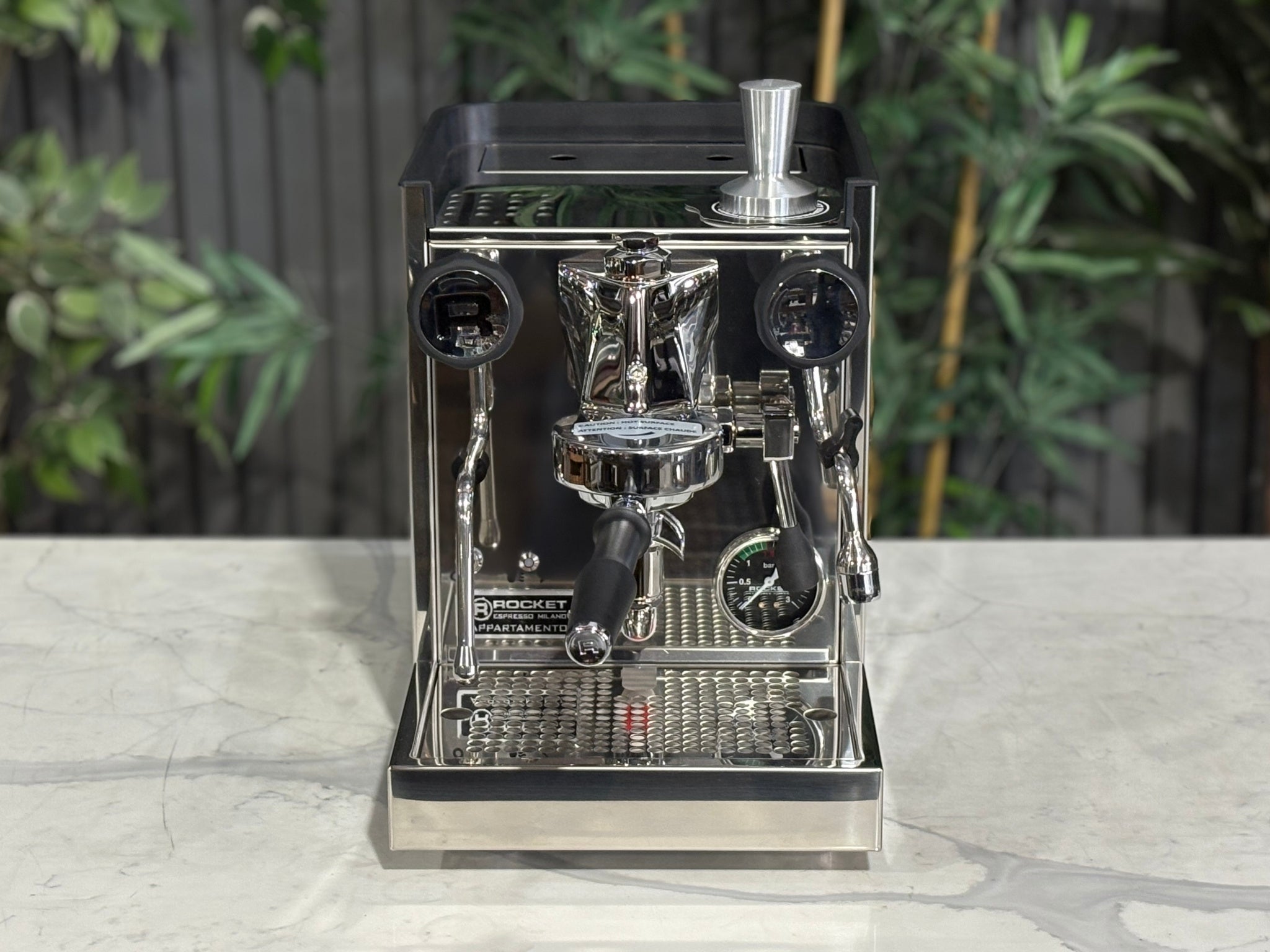Rocket Appartamento TCA 1 Group Coffee Machine Stainless with Black Cutouts - New, front-top perspective showing the drip tray, control lever, and E61 group head with stainless steel body and black cutouts on a marble bench with greenery in the background.