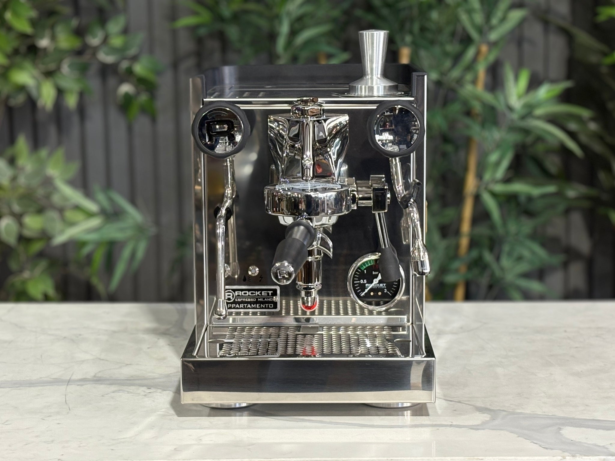 Rocket Appartamento TCA 1 Group Coffee Machine Stainless with Black Cutouts - New, front-on view highlighting the E61 group head, mechanical thermal control lever, high cup clearance, and drip tray on a marble bench with greenery in the background.