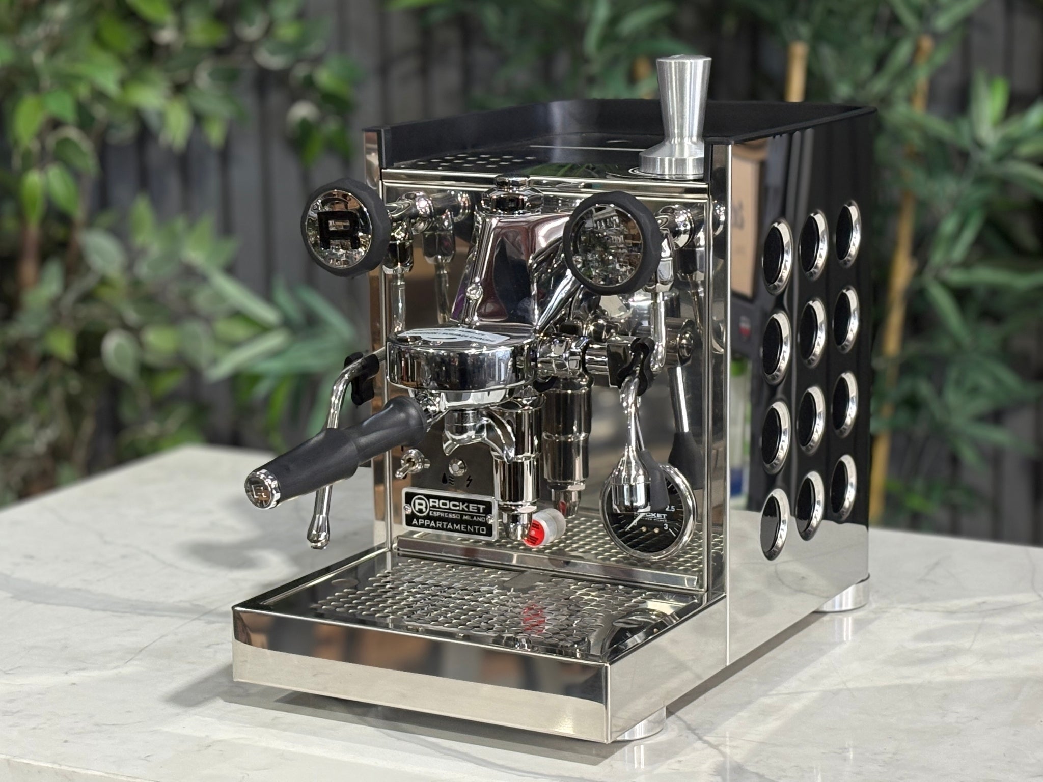 Rocket Appartamento TCA 1 Group Coffee Machine Stainless with Black Cutouts - New, angled view showing the E61 group head, high cup clearance, drip tray, and stainless steel body with black cutouts on a marble bench with greenery in the background.