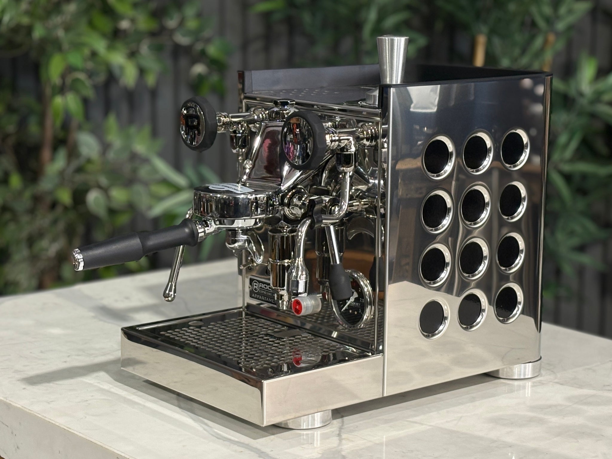 Rocket Appartamento TCA 1 Group Coffee Machine Stainless with Black Cutouts - New, angled view showing the E61 group head, high cup clearance, drip tray, and stainless steel body with black cutouts on a marble bench with greenery in the background.