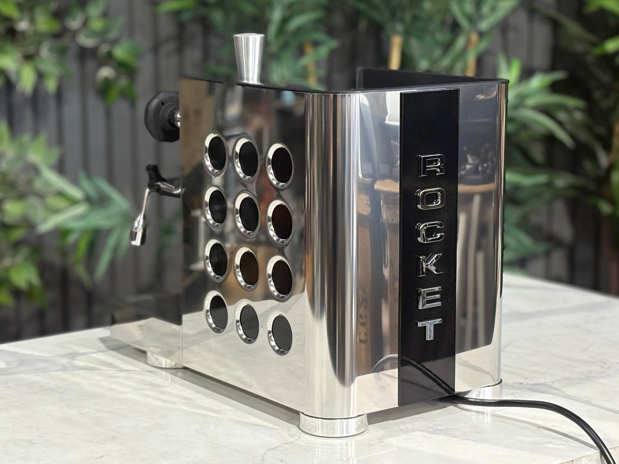 Rocket Appartamento TCA 1 Group Coffee Machine Stainless with Black Cutouts - New, angled rear view showing the vertical Rocket logo, black cutouts, and rounded stainless steel edges on a marble bench with greenery in the background.