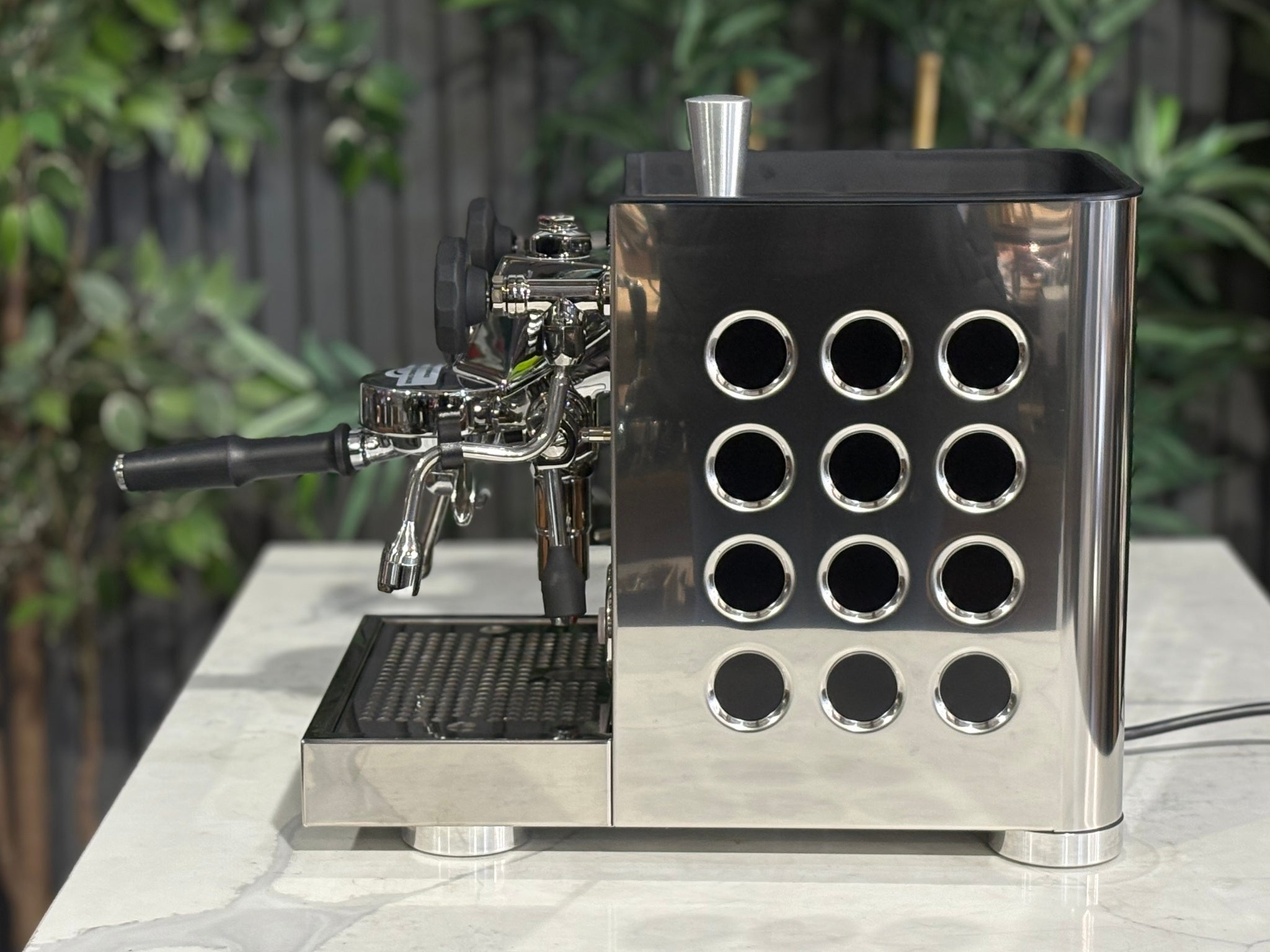 Rocket Appartamento TCA 1 Group Coffee Machine Stainless with Black Cutouts - New, side view displaying the circular debossed panel, rounded edges, and stainless steel body with black cutouts on a marble bench with greenery in the background.